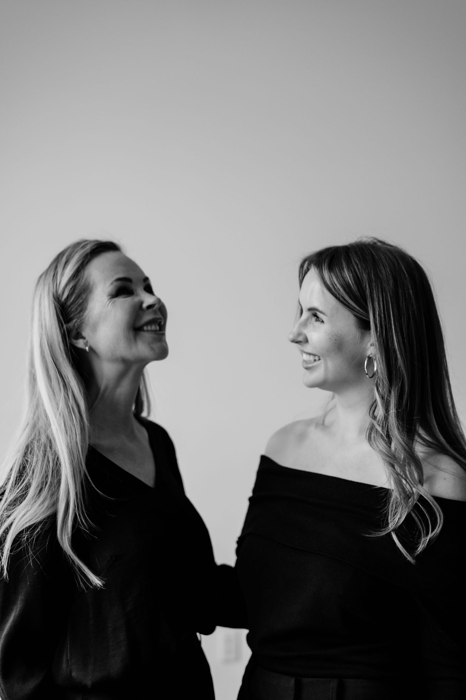 Two women in black tops smile at each other in a stylish black and white portrait photo.
