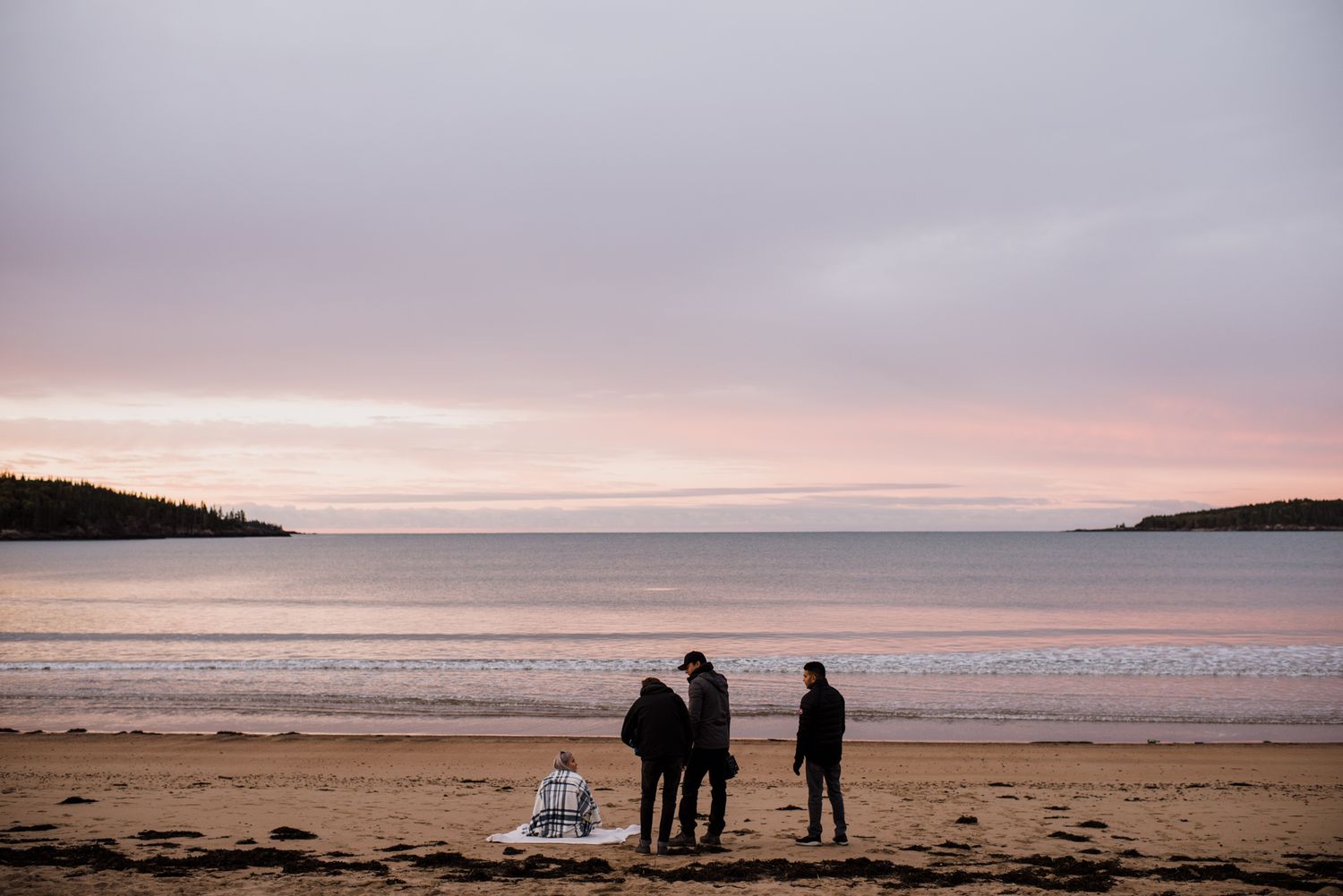 Three people standing on a sandy beach at sunset with pink and purple sky over calm sea.
