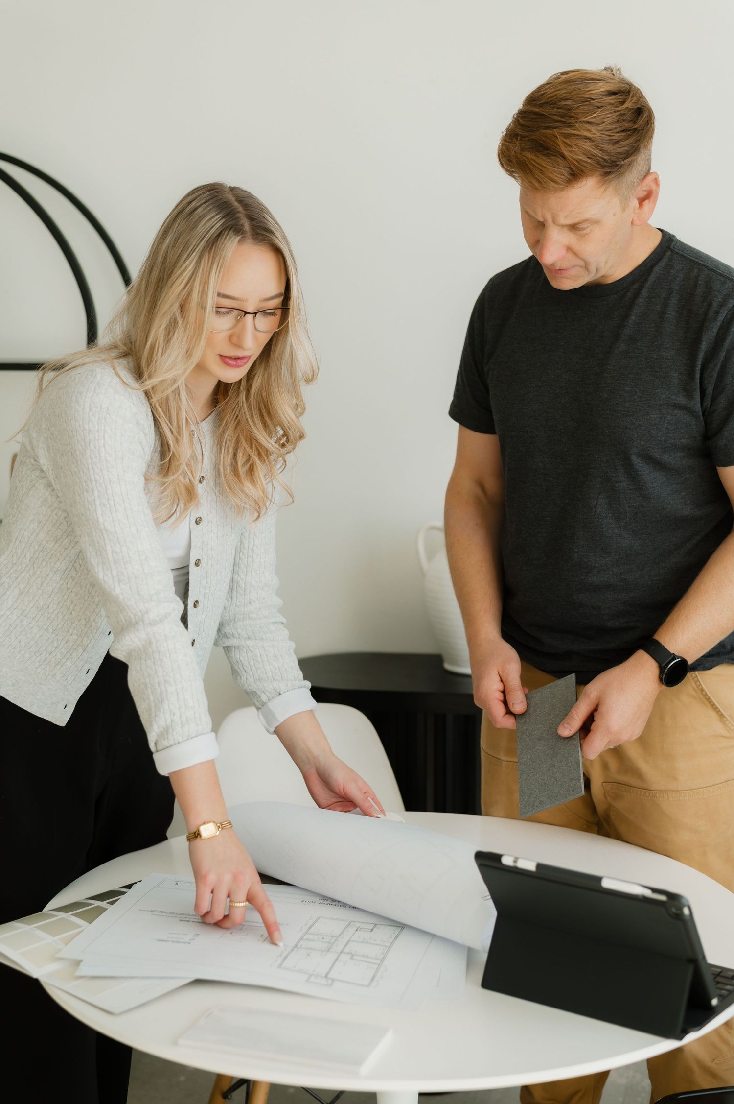 Two colleagues review architectural blueprints together at a round white table in a bright office.