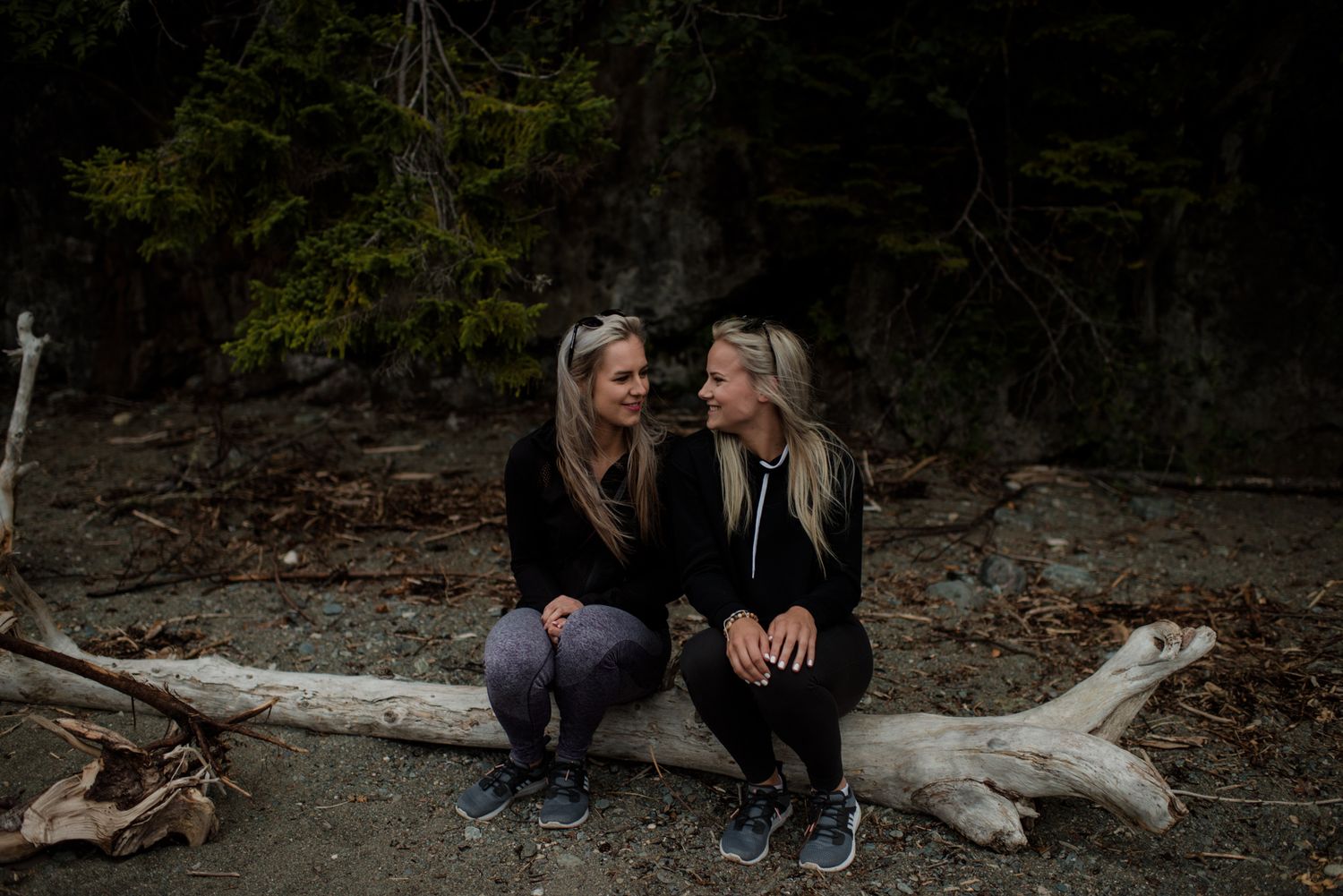 Two blonde women sitting together on a driftwood log on a dark forest beach.