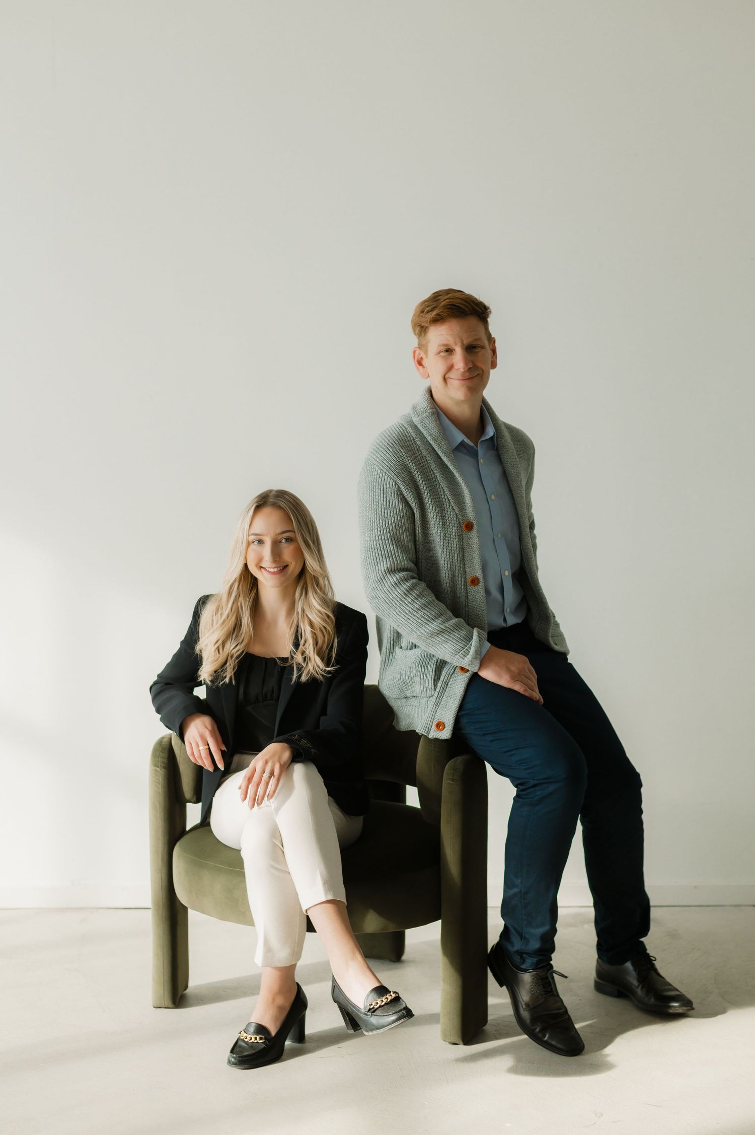 Two smiling professionals, a blonde woman in black and a man in gray cardigan, pose on modern chairs against a white wall.