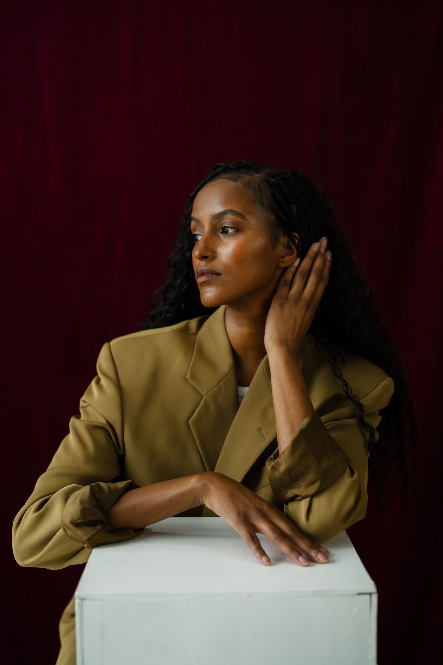 Woman in olive blazer leaning on white cube, gazing sideways against deep red backdrop.