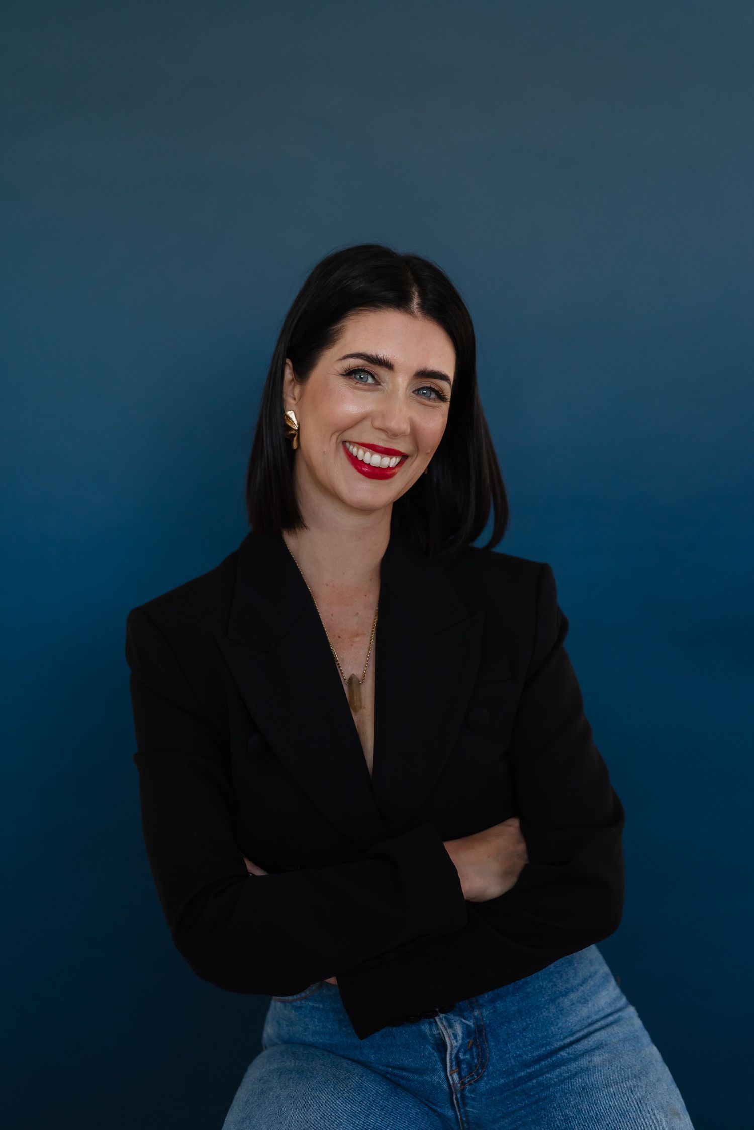A person in a black blazer and jeans poses against a dark blue background with a bright smile.