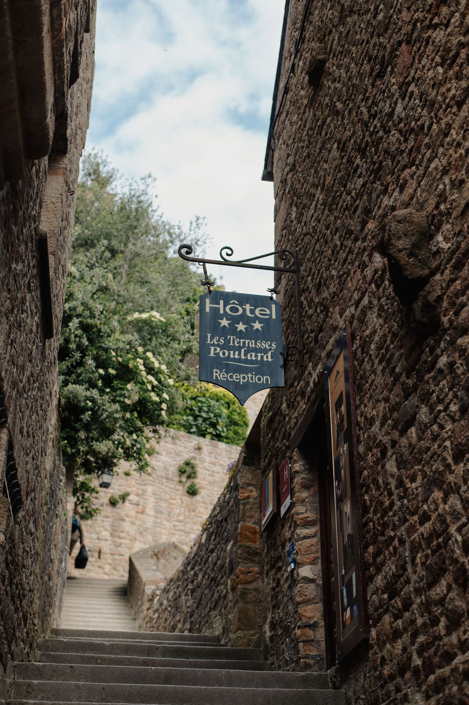 A narrow stone alleyway leads to Hotel Les Terrasses Poulard reception sign in Mont Saint-Michel, France.