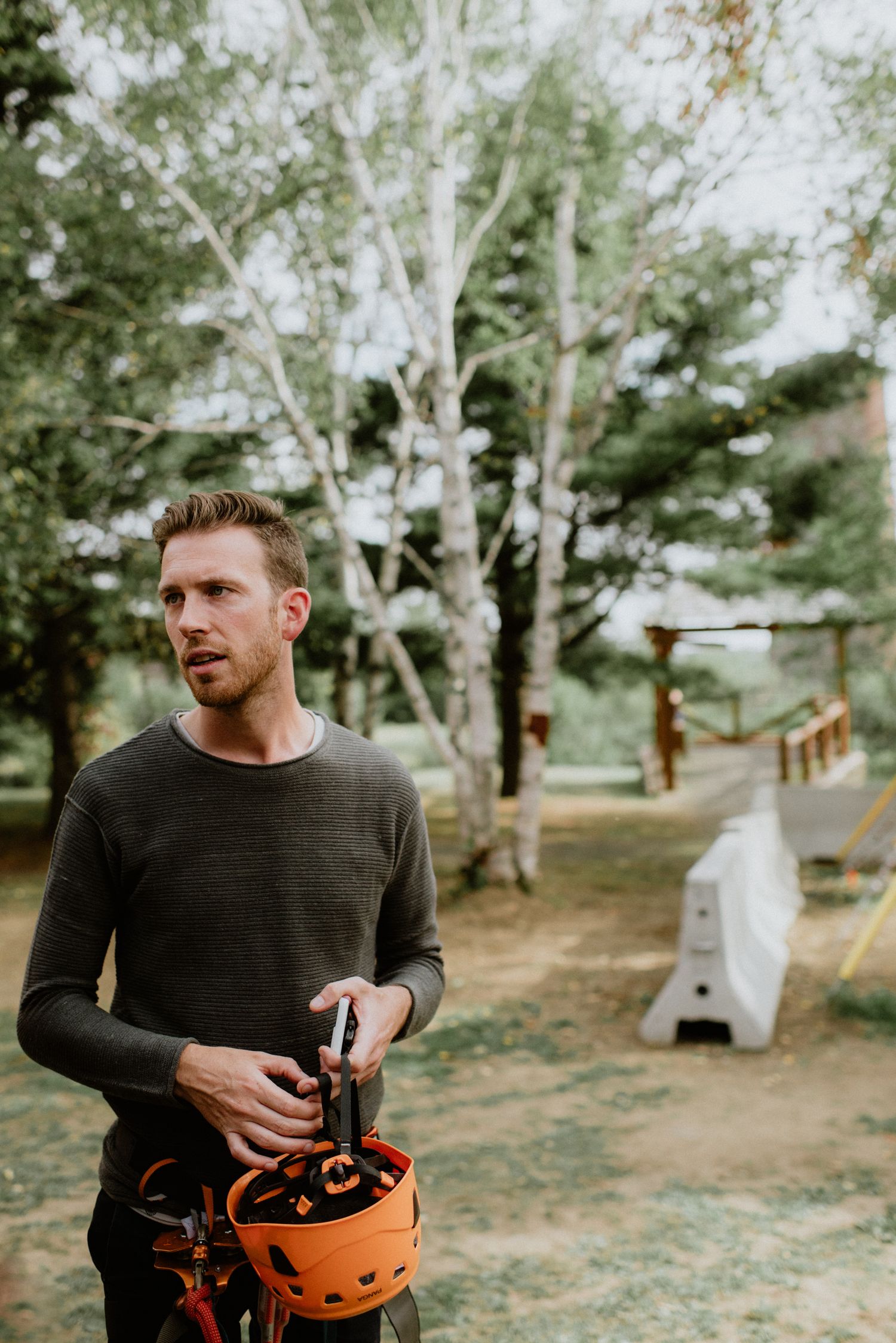A man in a dark sweater holds an orange helmet outdoors near trees at an adventure park.