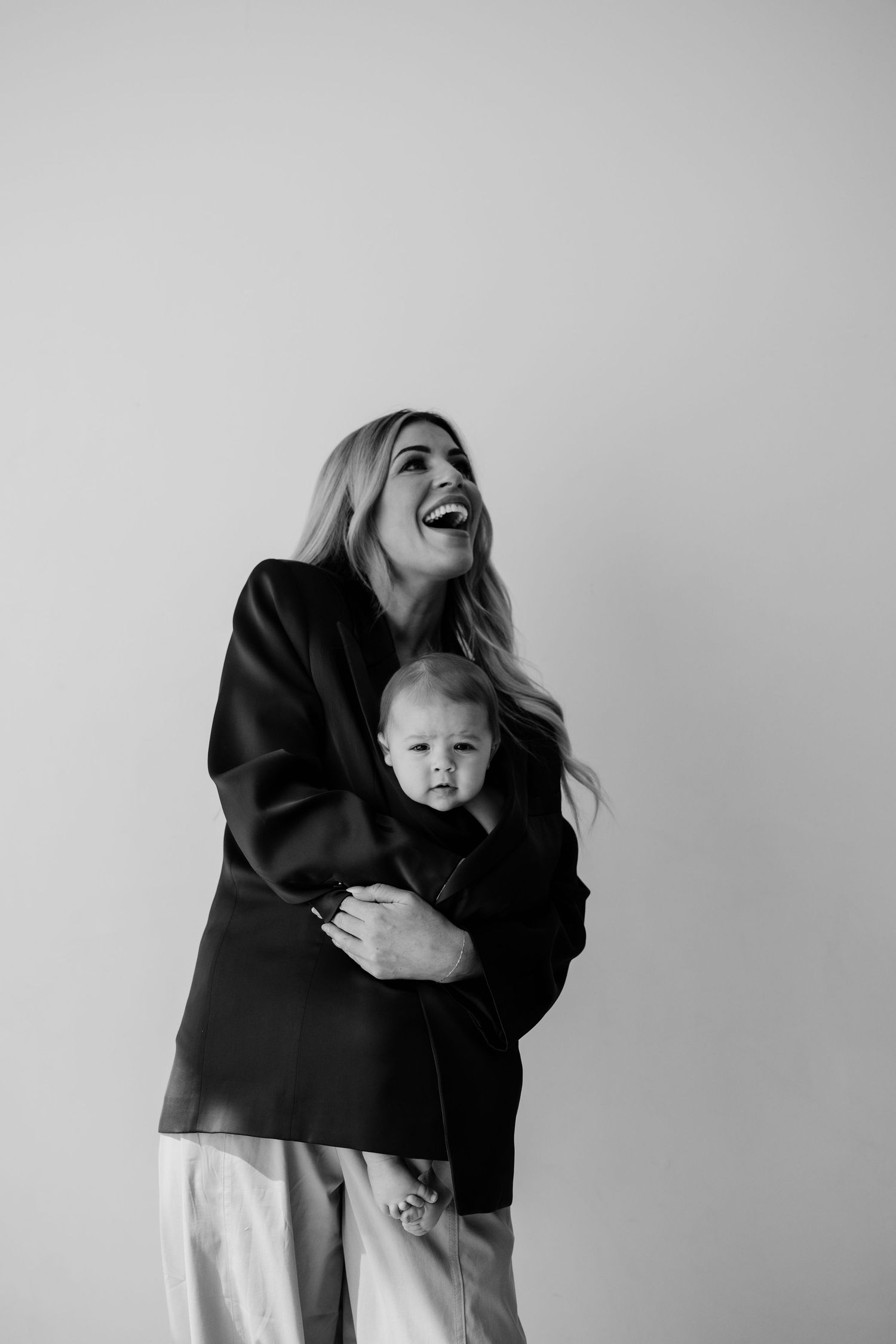 Black and white photo of a laughing woman in a blazer holding a baby against a light background.