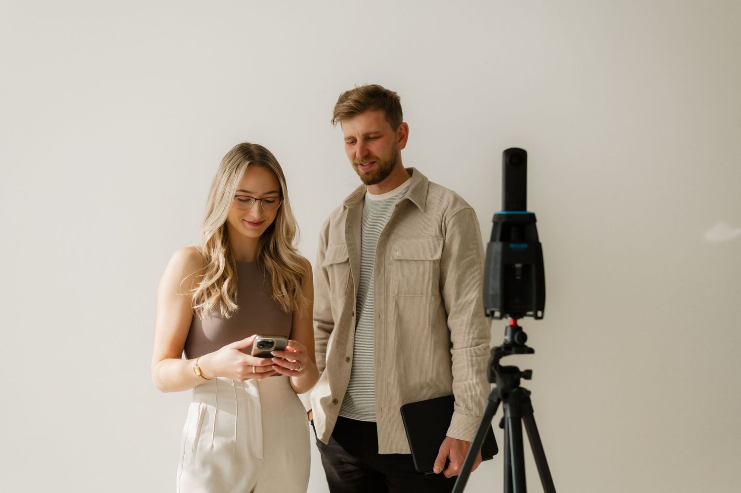 A man and woman stand together reviewing content on a smartphone next to a camera on a tripod.