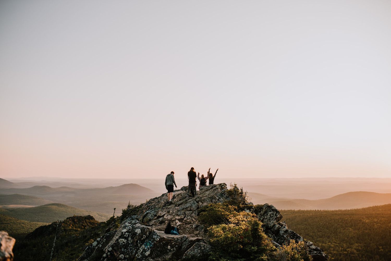 Group of hikers celebrating on a rocky mountain summit at sunset with vast landscape views.