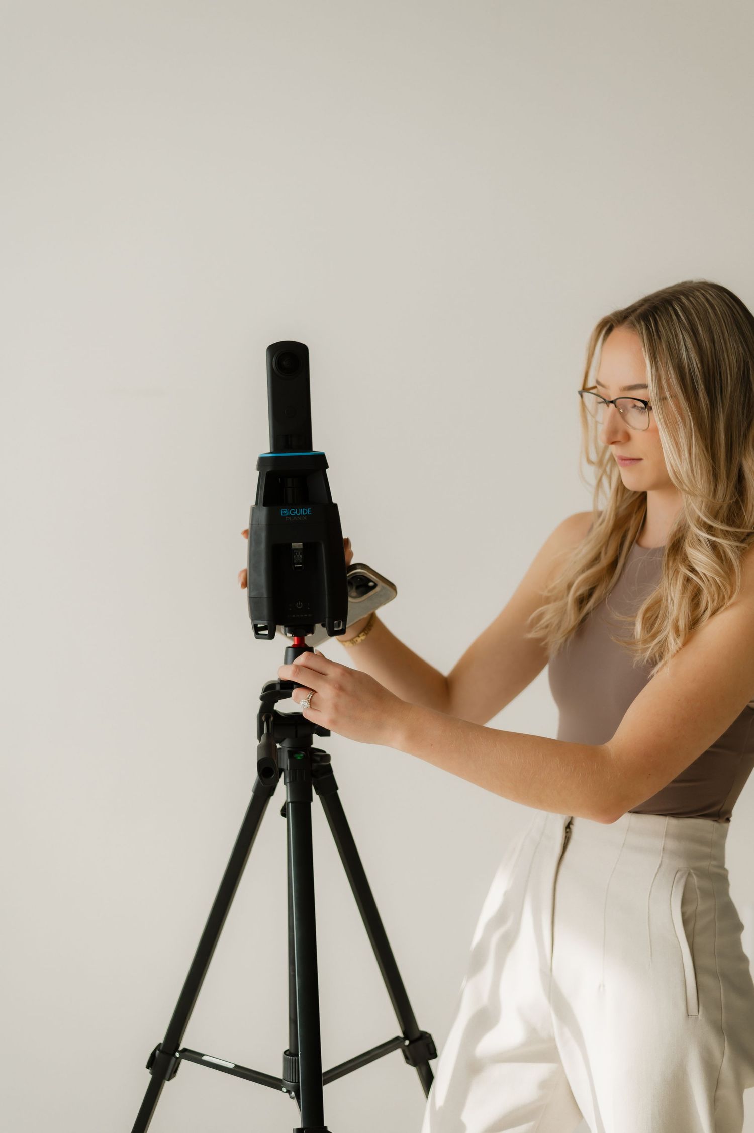 Woman with glasses adjusting a camera on a tripod against a white background in a bright studio setting.