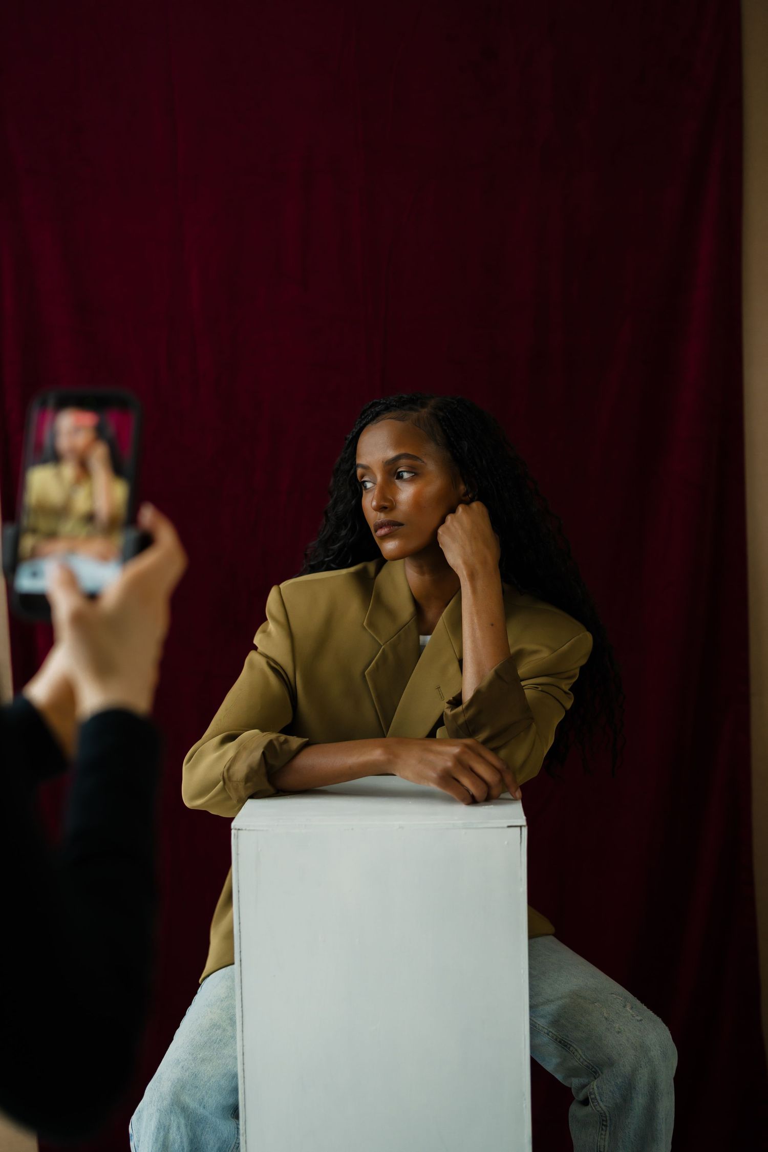 Woman in olive blazer poses on white cube while being photographed against dark red curtain backdrop.