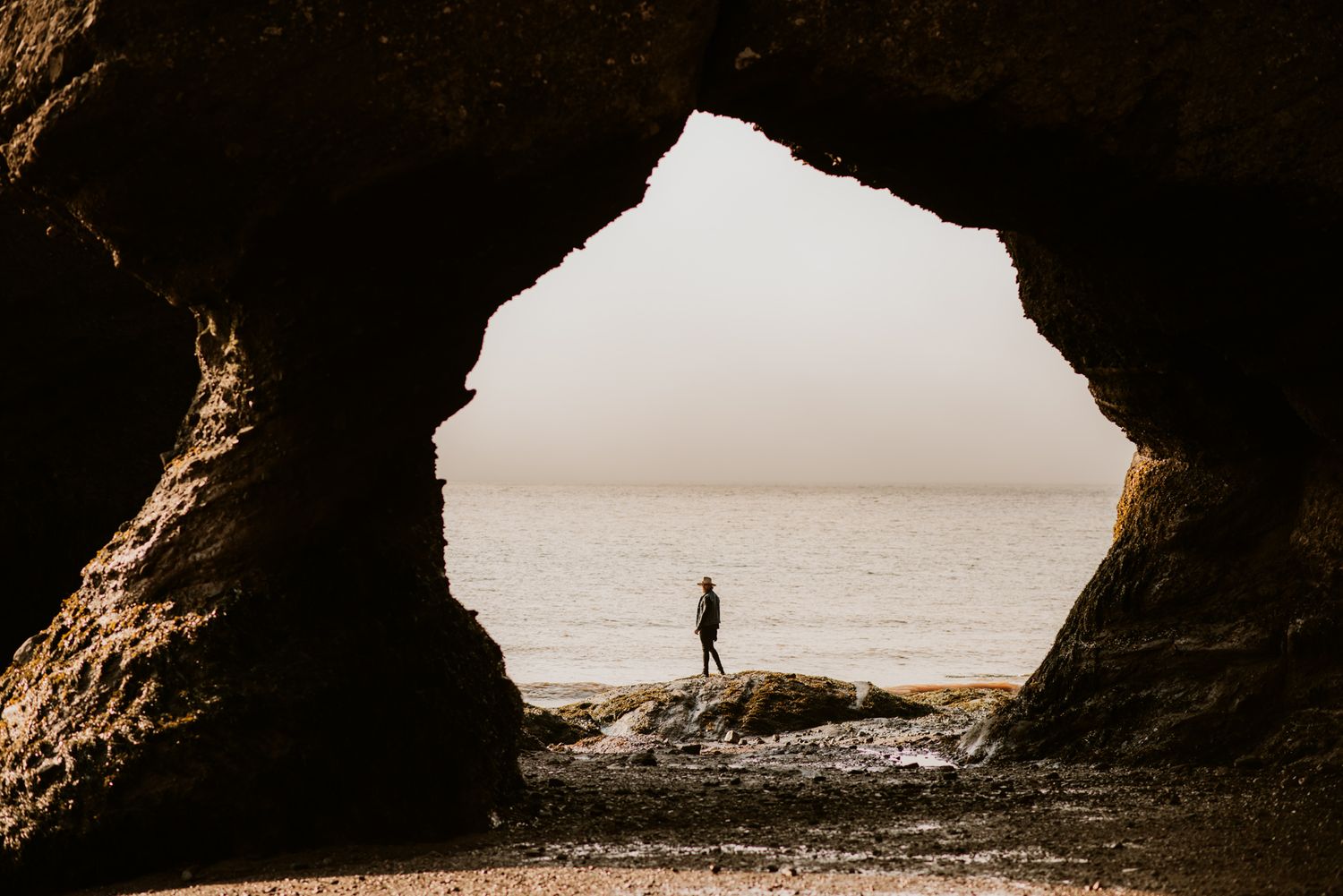 Silhouette of a person standing on a rocky beach, framed by a dramatic sea cave opening with ocean views.