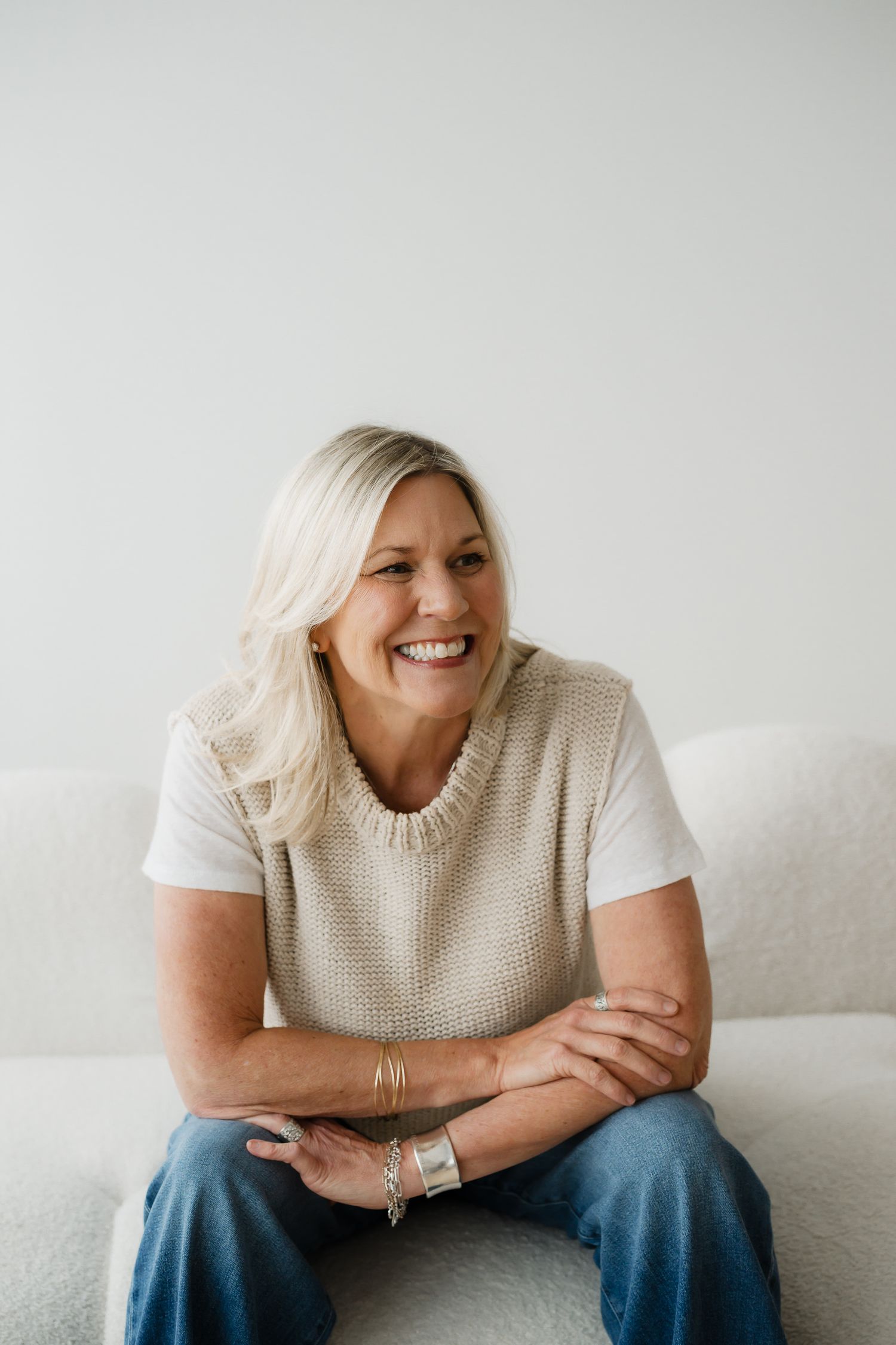 Smiling blonde woman in beige knit vest and jeans sitting on white sofa with arms crossed.