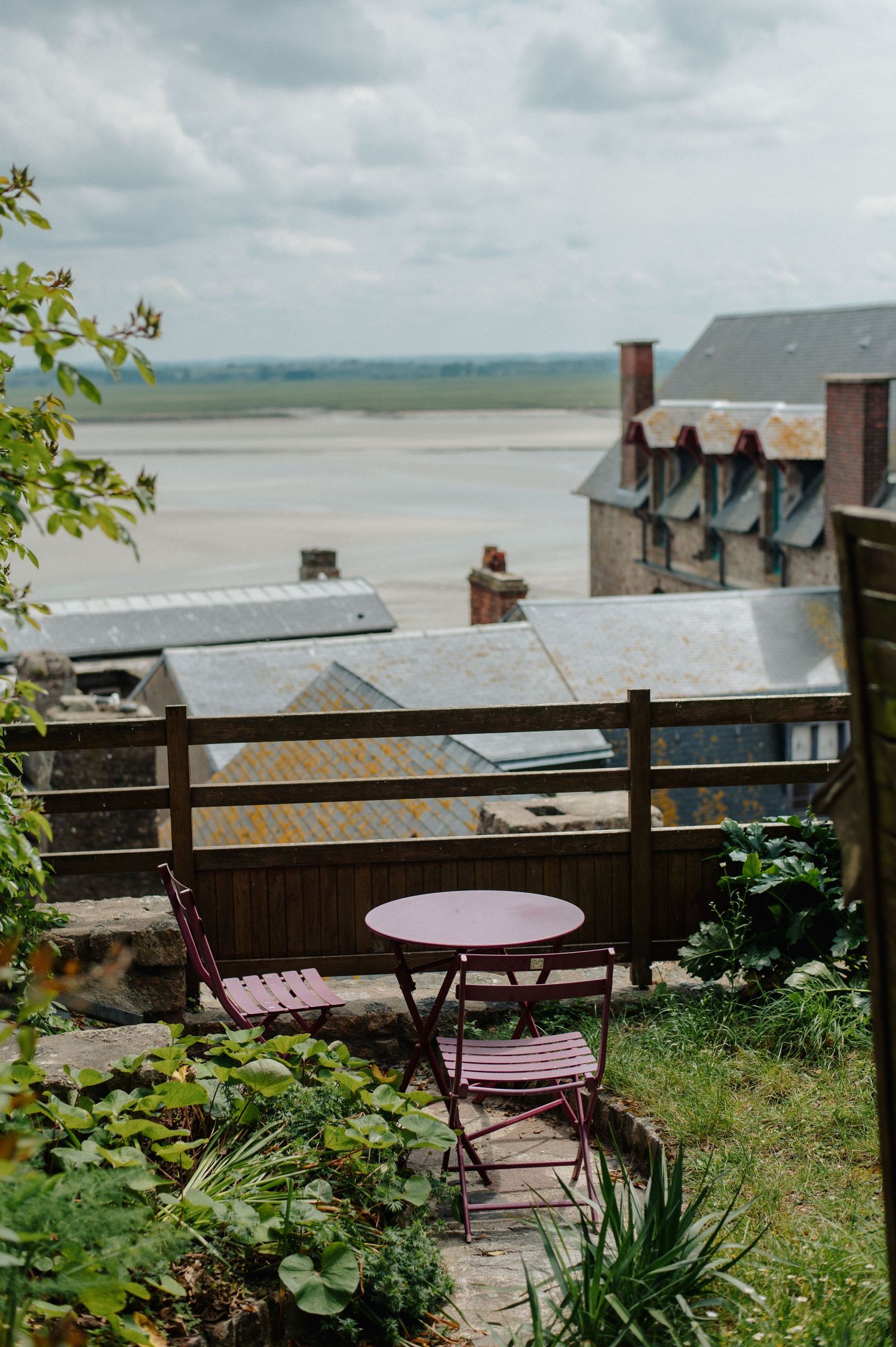 Pink bistro table and chairs on a terrace overlooking tidal flats and rooftops under cloudy skies.