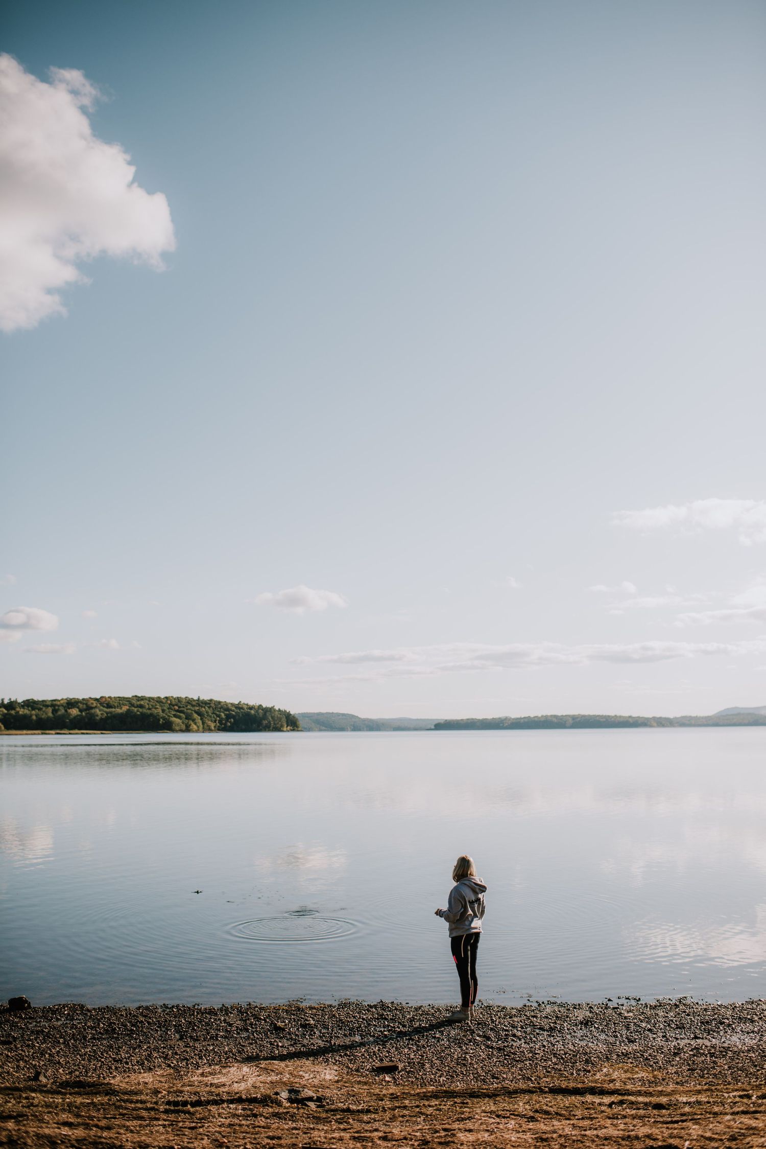 Person standing at the edge of a calm lake shore under a partly cloudy sky with distant tree-lined hills.