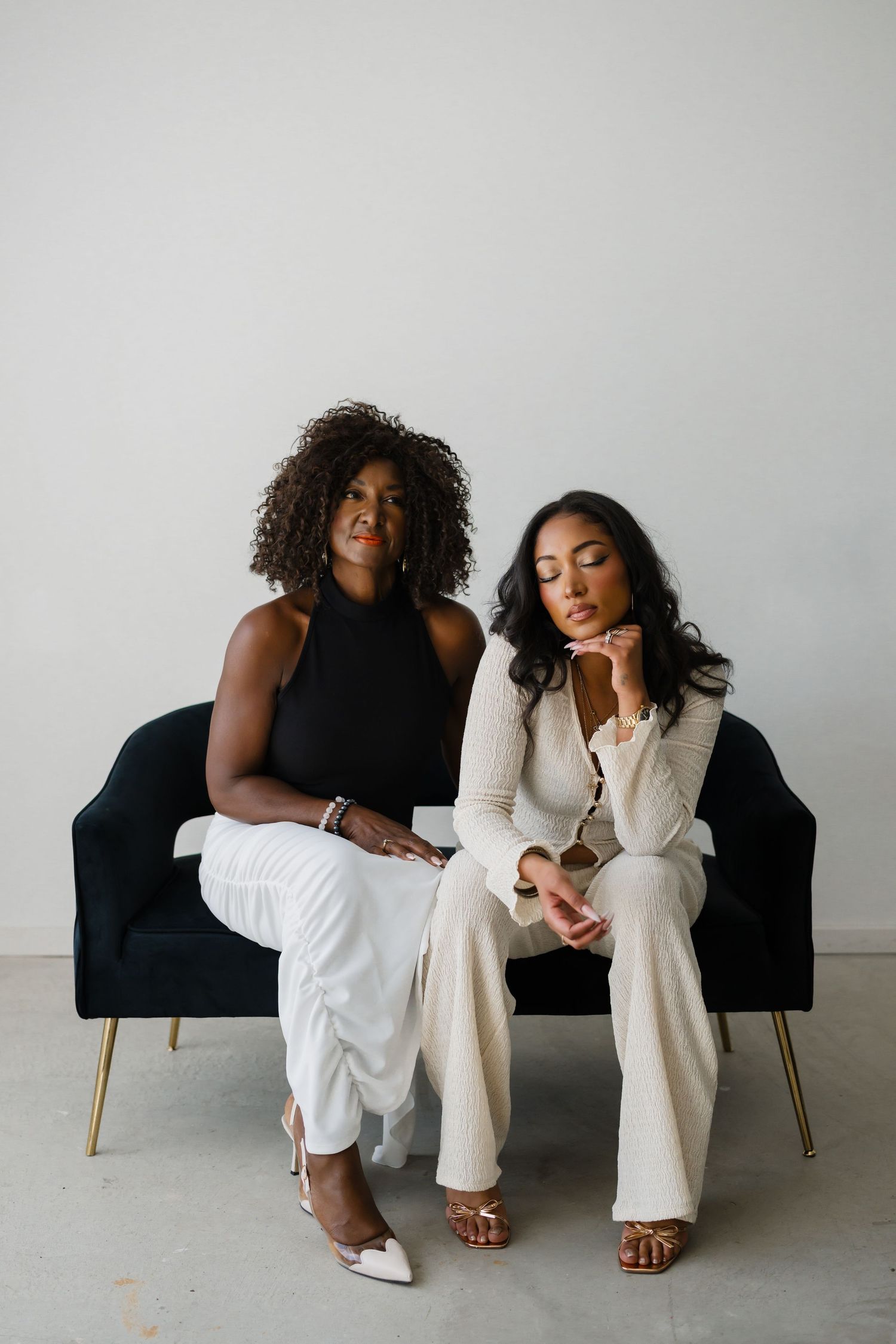 Two stylish women sitting on a black velvet chair, wearing white and beige outfits against a white background.