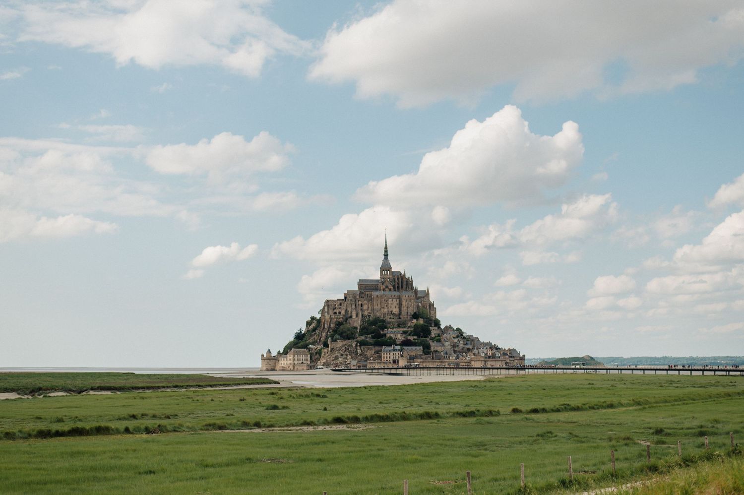 Mont Saint-Michel island abbey rising above green meadows under a partly cloudy sky in Normandy, France.