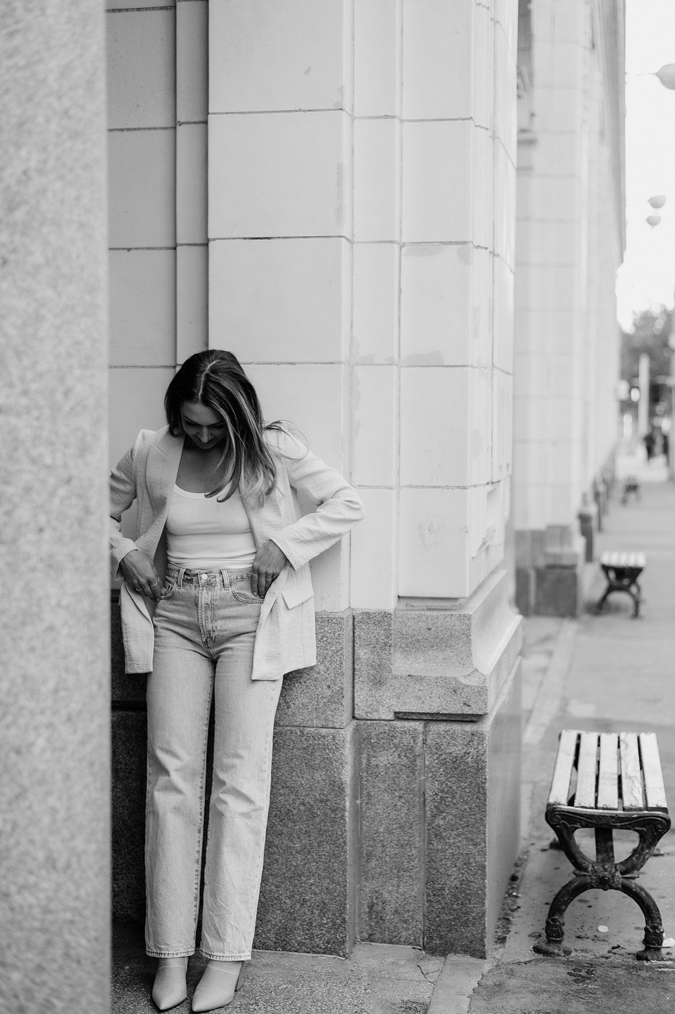 Black and white photo of a stylish woman in blazer and jeans leaning against a stone building column on a city street.