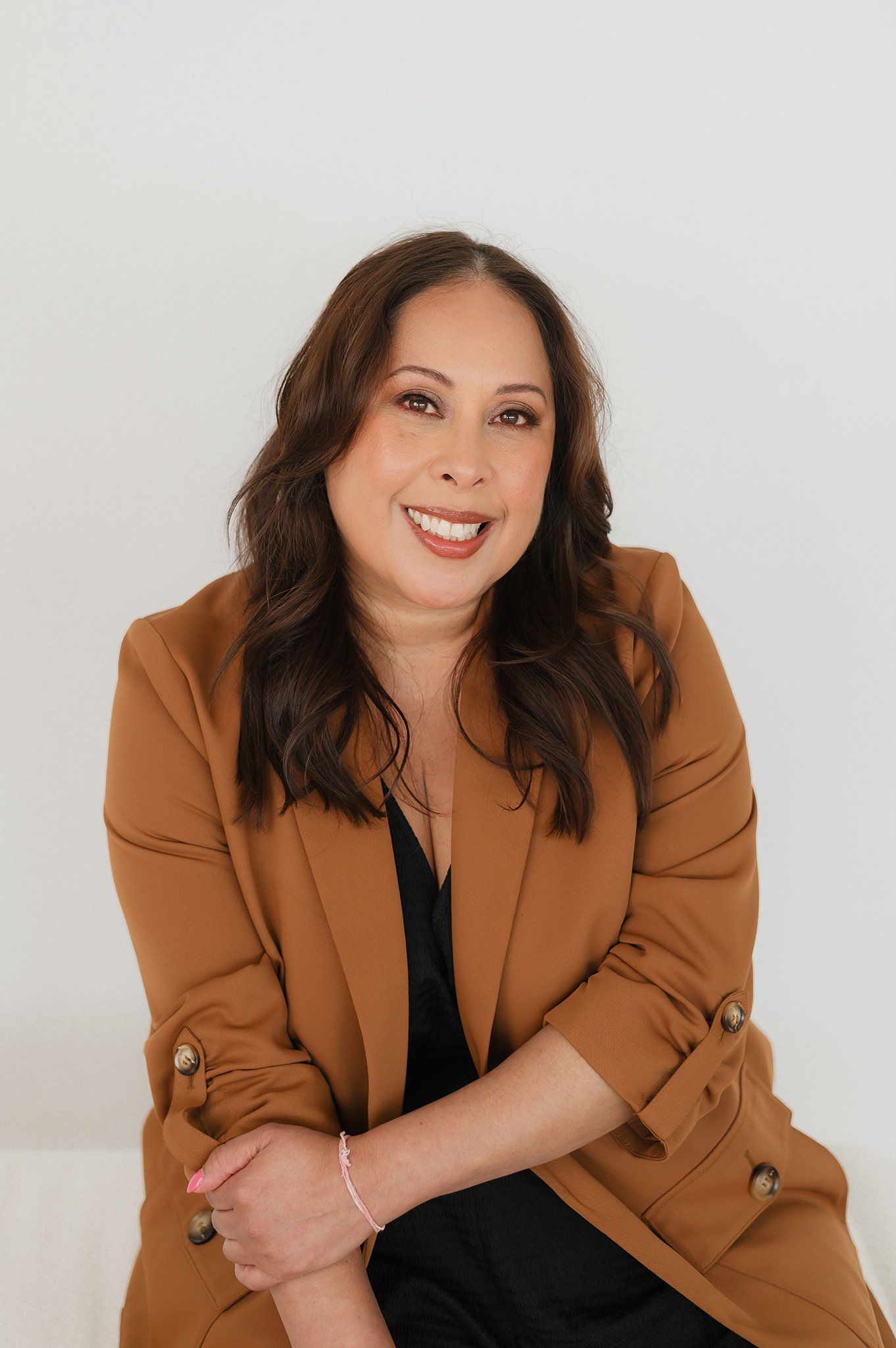 Professional portrait of someone wearing a camel-colored blazer over black top against white background, smiling warmly.