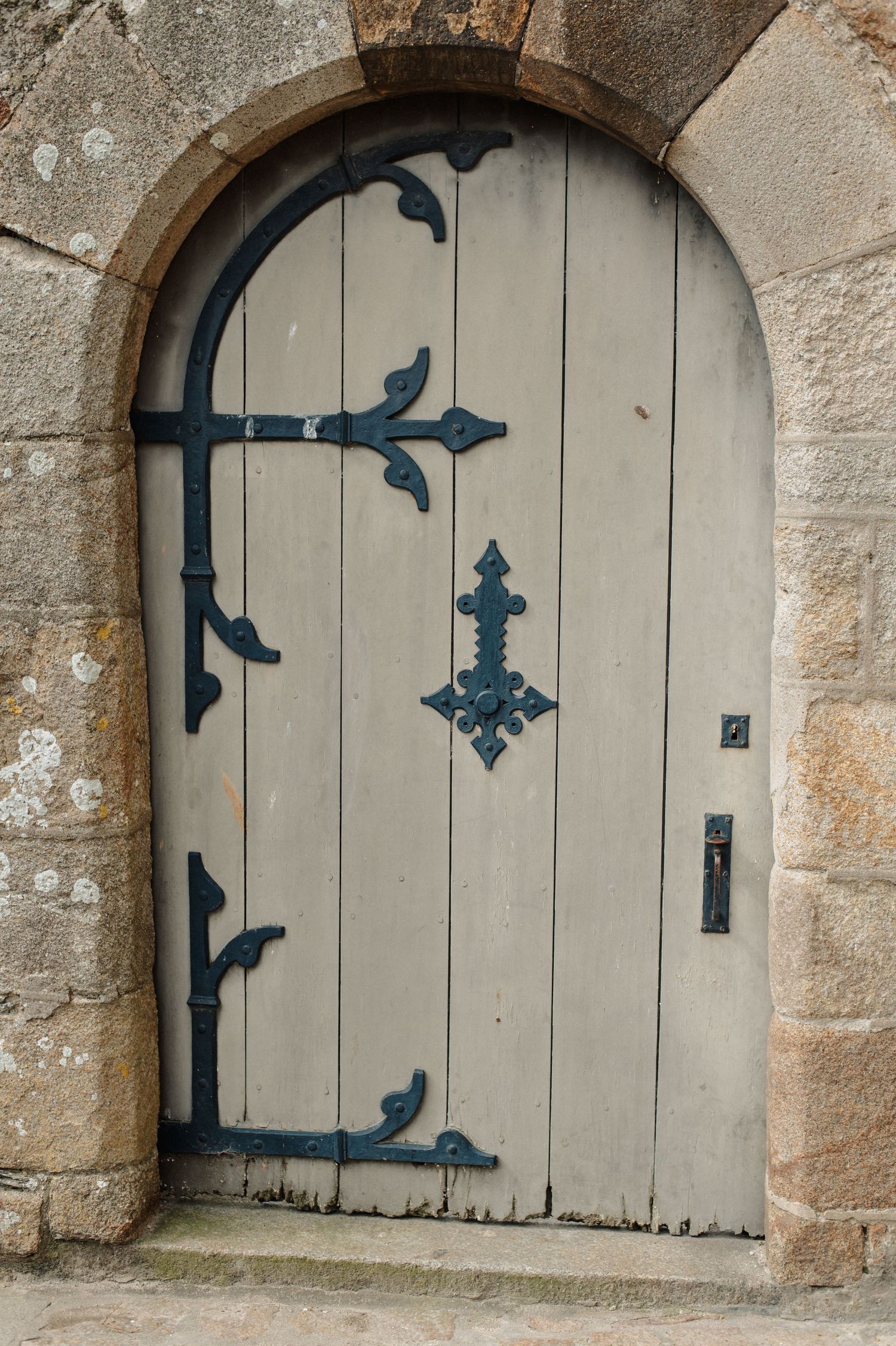 Antique arched wooden door with ornate black iron hinges and hardware set in a stone wall.