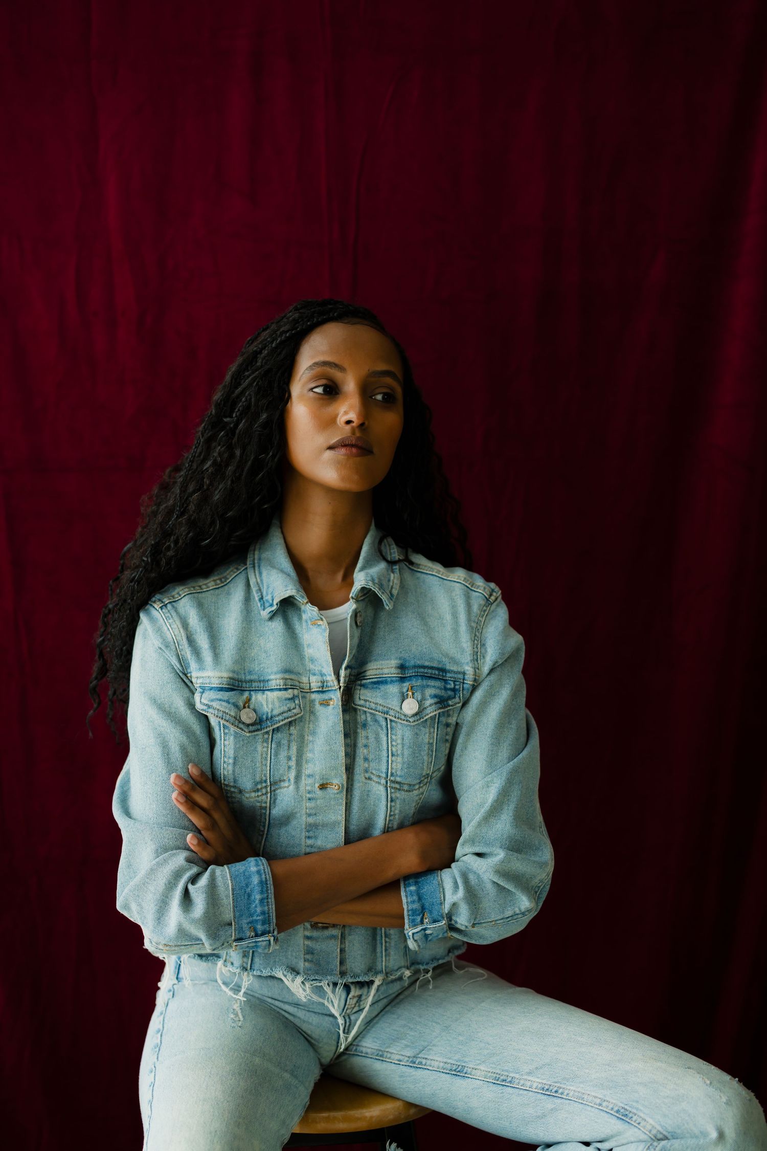 Young woman in denim jacket sitting with arms crossed against deep red fabric background.