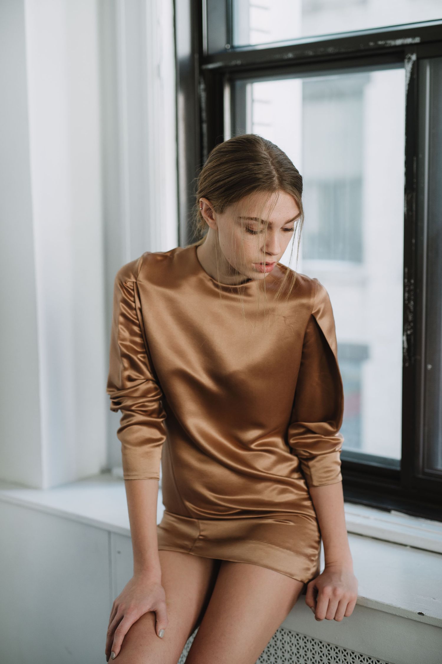 Woman in golden satin mini dress sitting by window, elegant ruched long sleeves, chic bronze silk outfit.