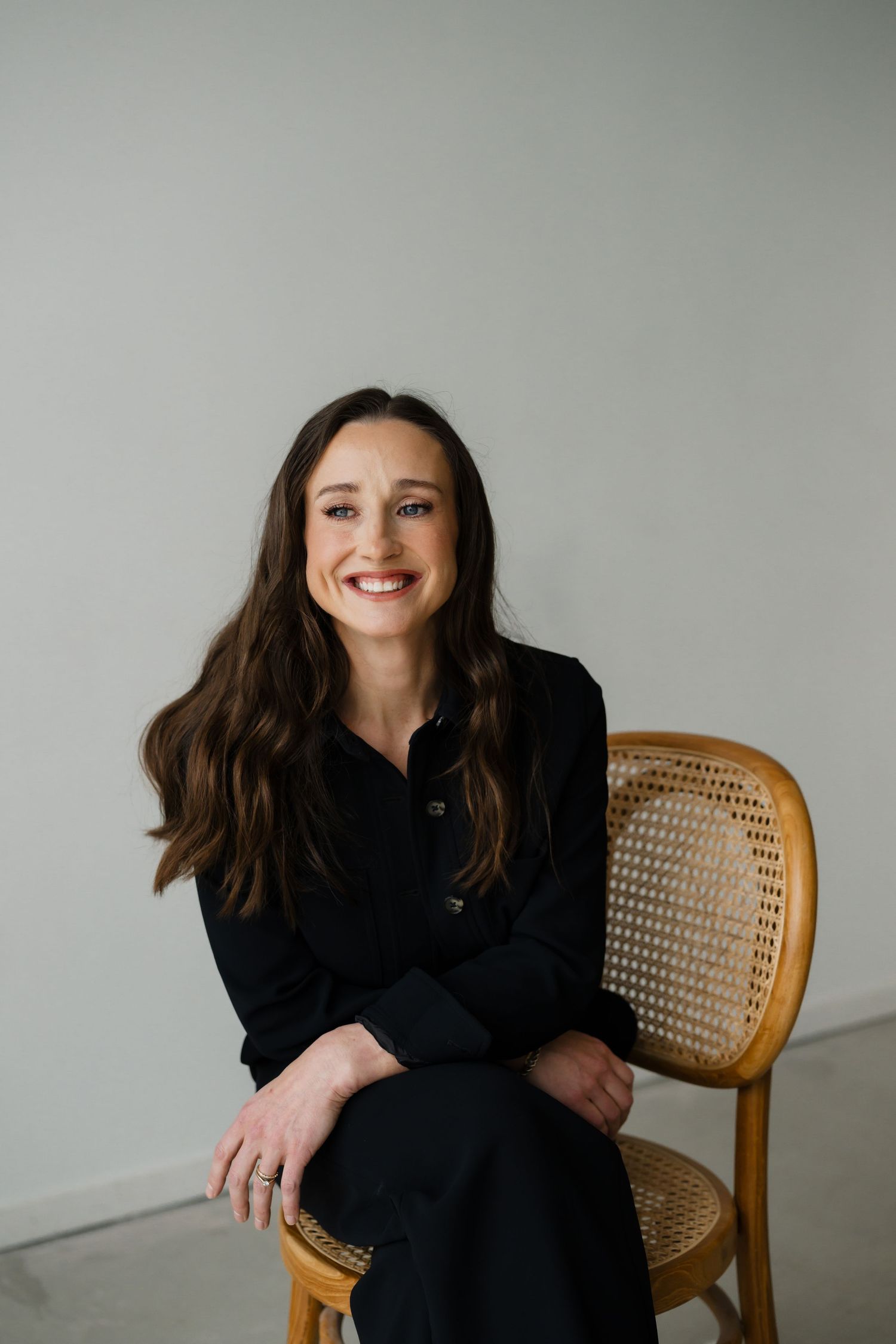 Smiling woman with long brown hair sitting on a wooden cane chair, wearing a black outfit against a gray wall.