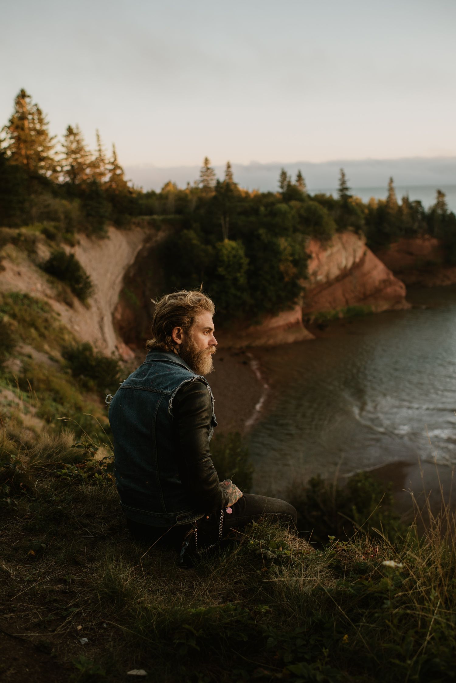 A bearded man in a leather jacket sits on a cliff edge overlooking a rocky coastal bay surrounded by evergreen trees.