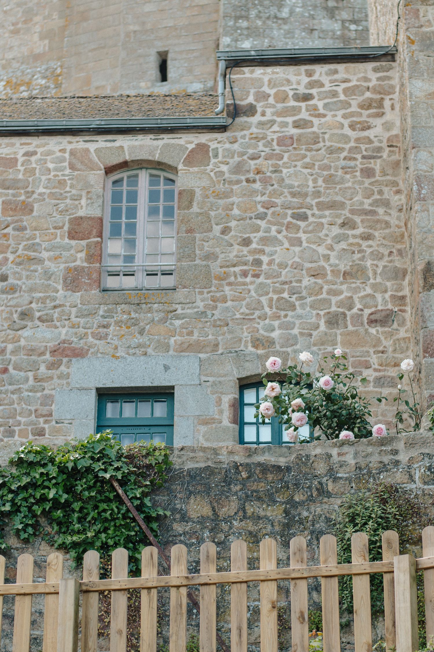 Historic stone building with green shuttered windows, climbing roses, ivy, and a wooden picket fence in a French village.