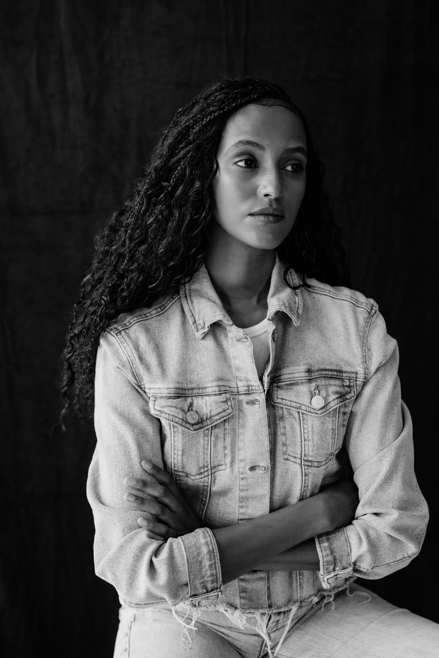 Black and white portrait of woman in denim jacket with arms crossed against dark background.