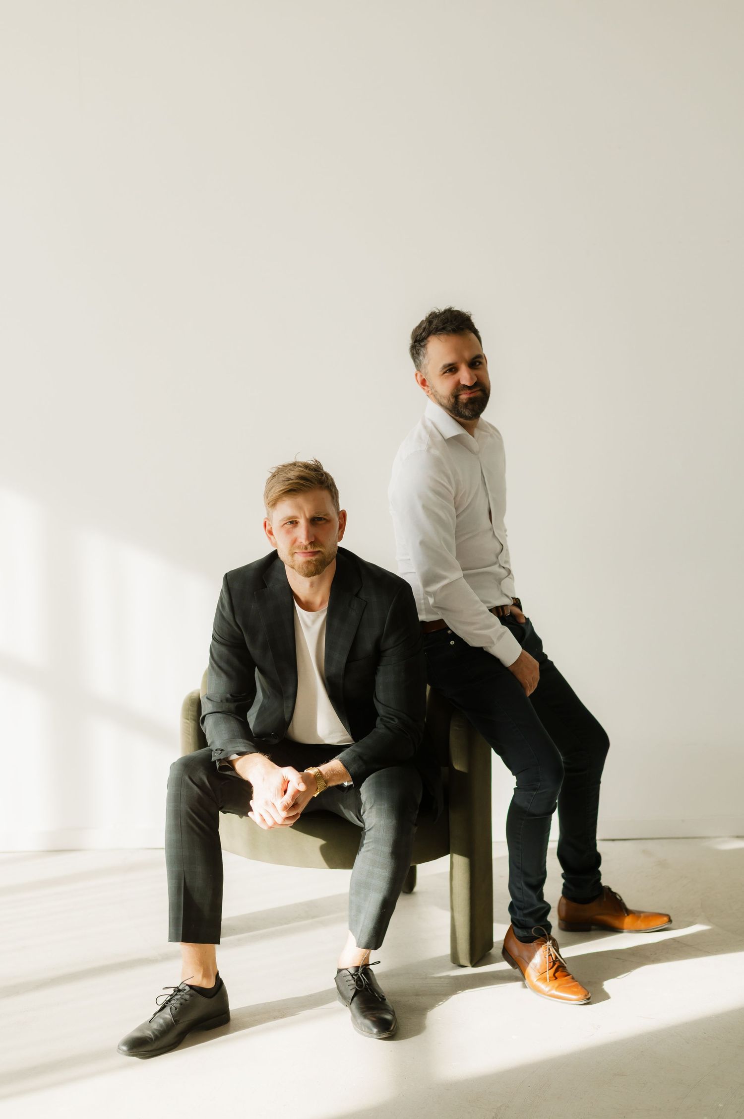 Two stylish men pose in a bright studio, one seated in a chair wearing a dark suit, the other standing in a white shirt.
