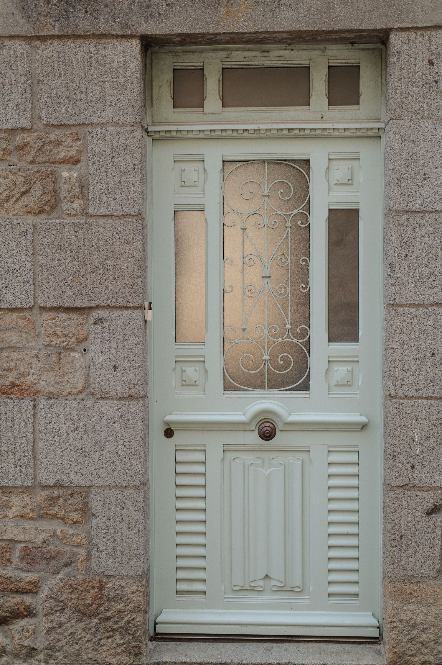 Elegant white painted wooden door with ornate iron scrollwork glass panel and louvered lower section set in stone wall.