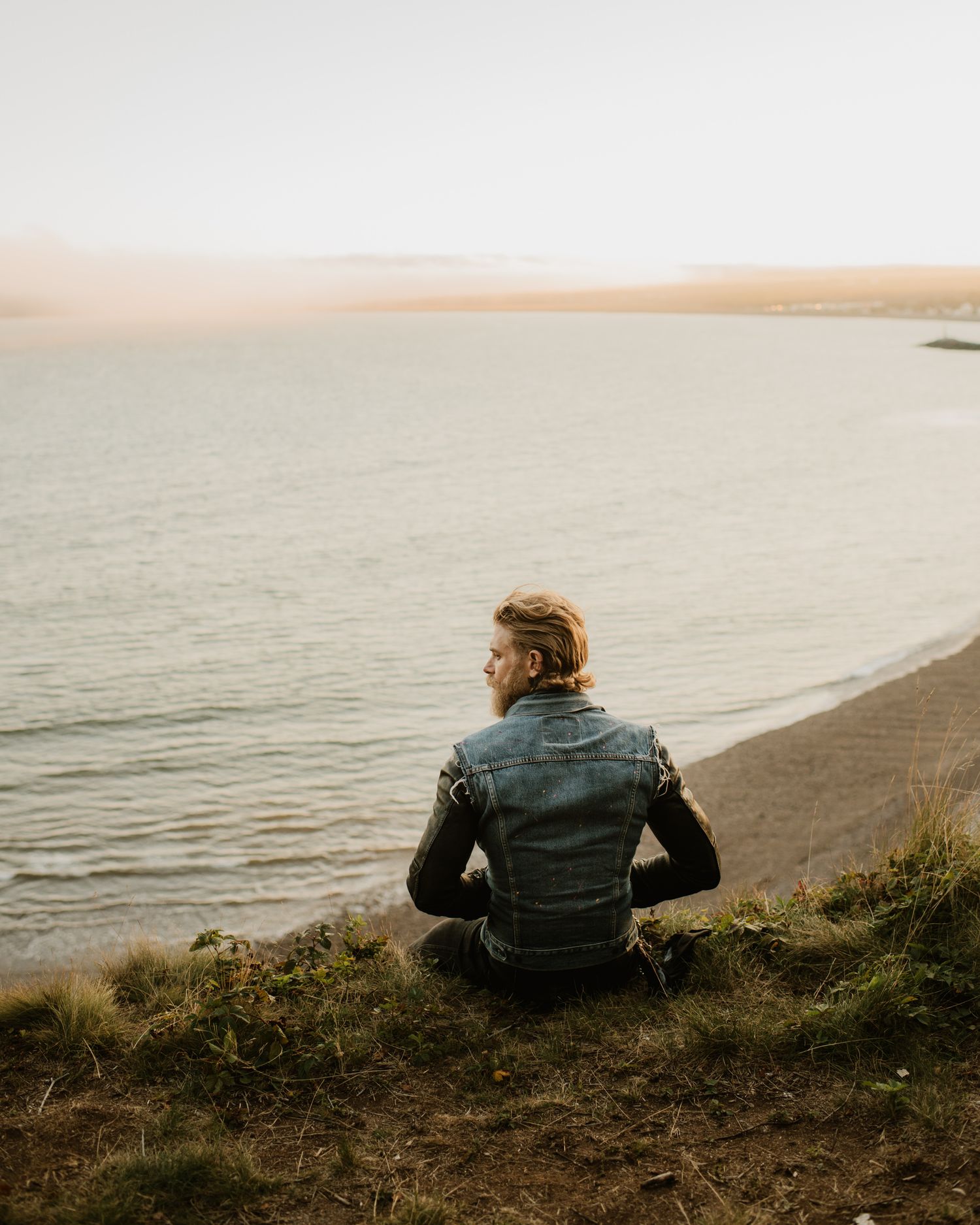 A man in a denim vest sits on a grassy cliff overlooking a calm lake shore at sunset.