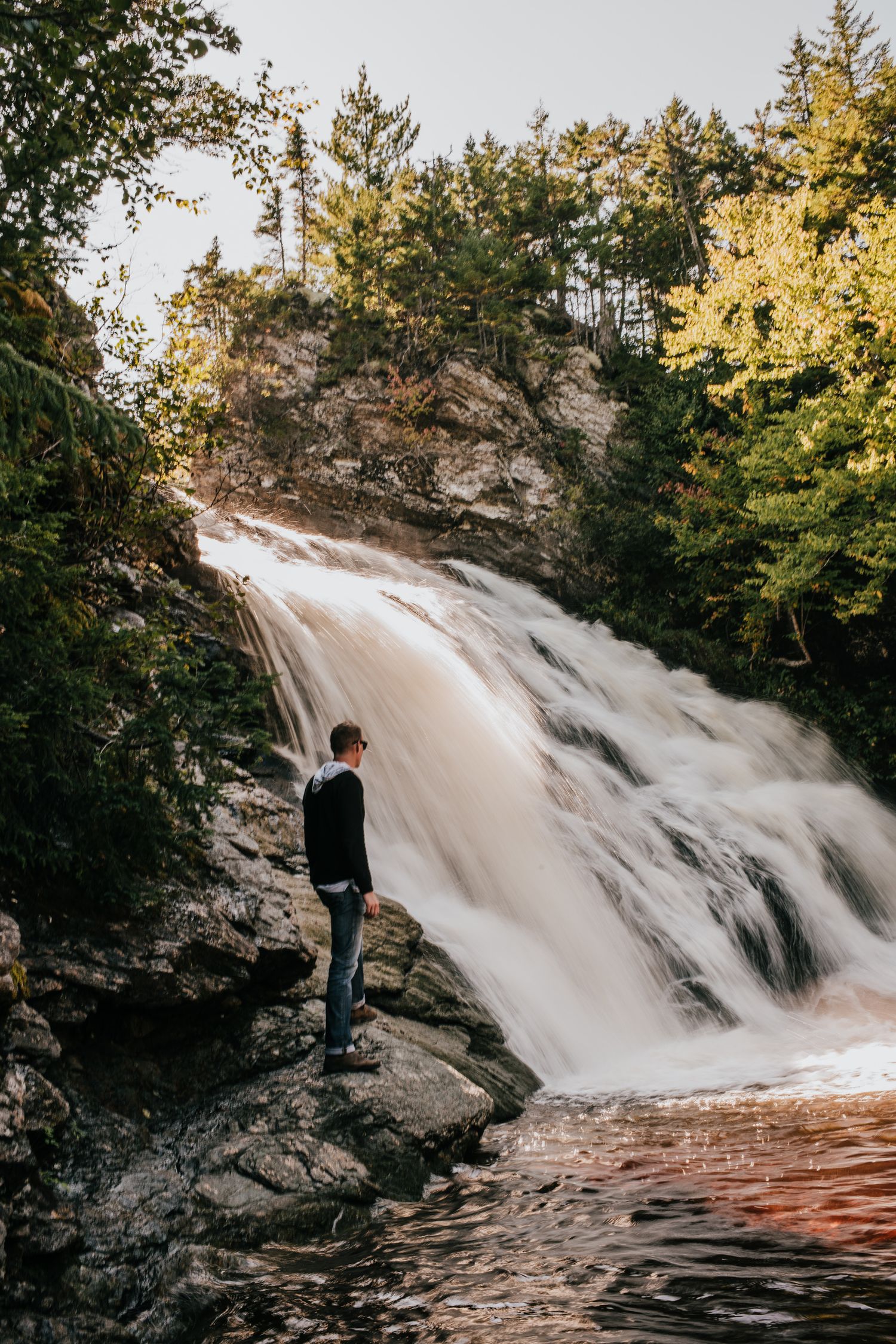 Person standing on rocks admiring a stunning cascading waterfall surrounded by lush green trees in a forest setting.