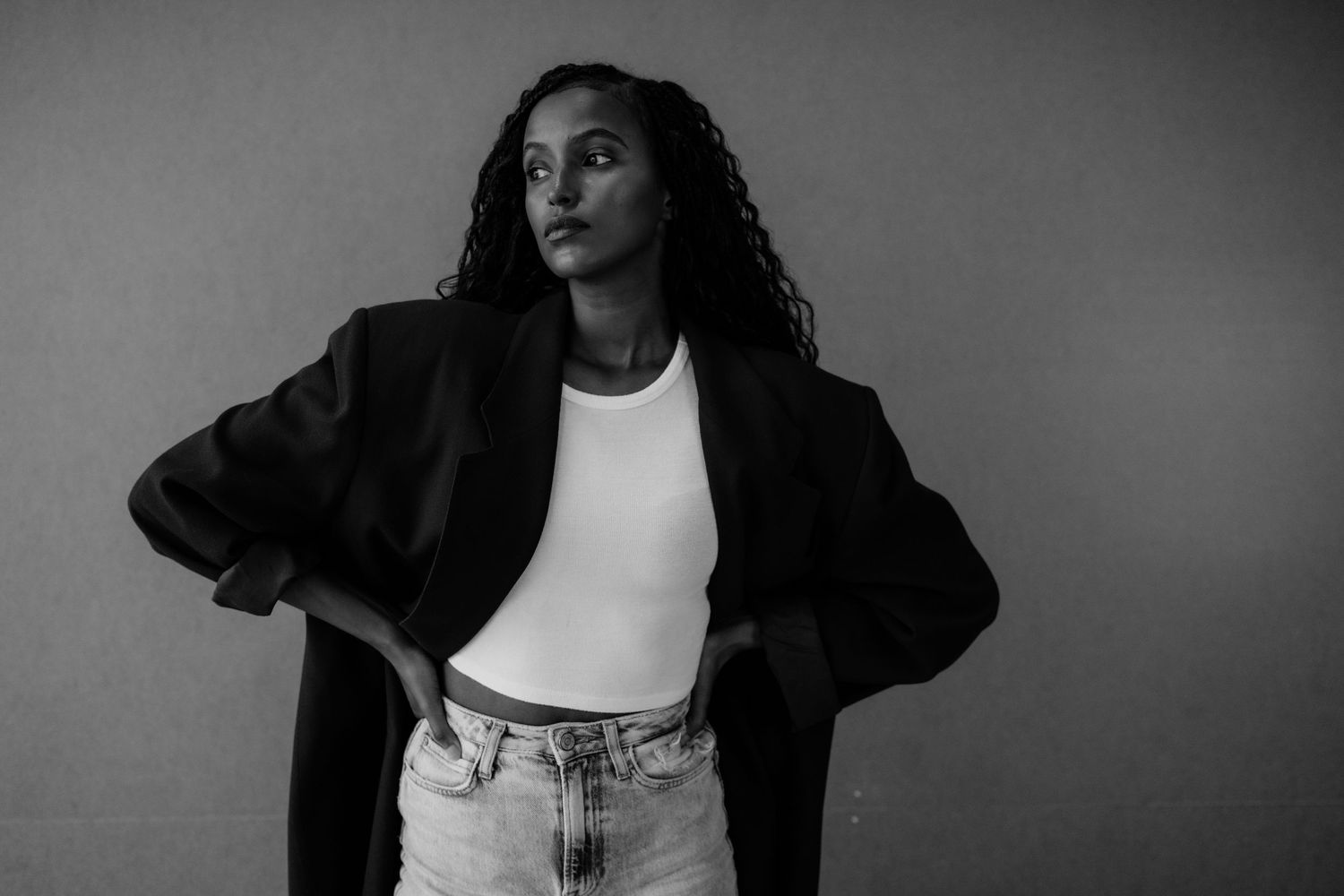 Black and white photo of woman in black blazer and jeans posing against gray background.
