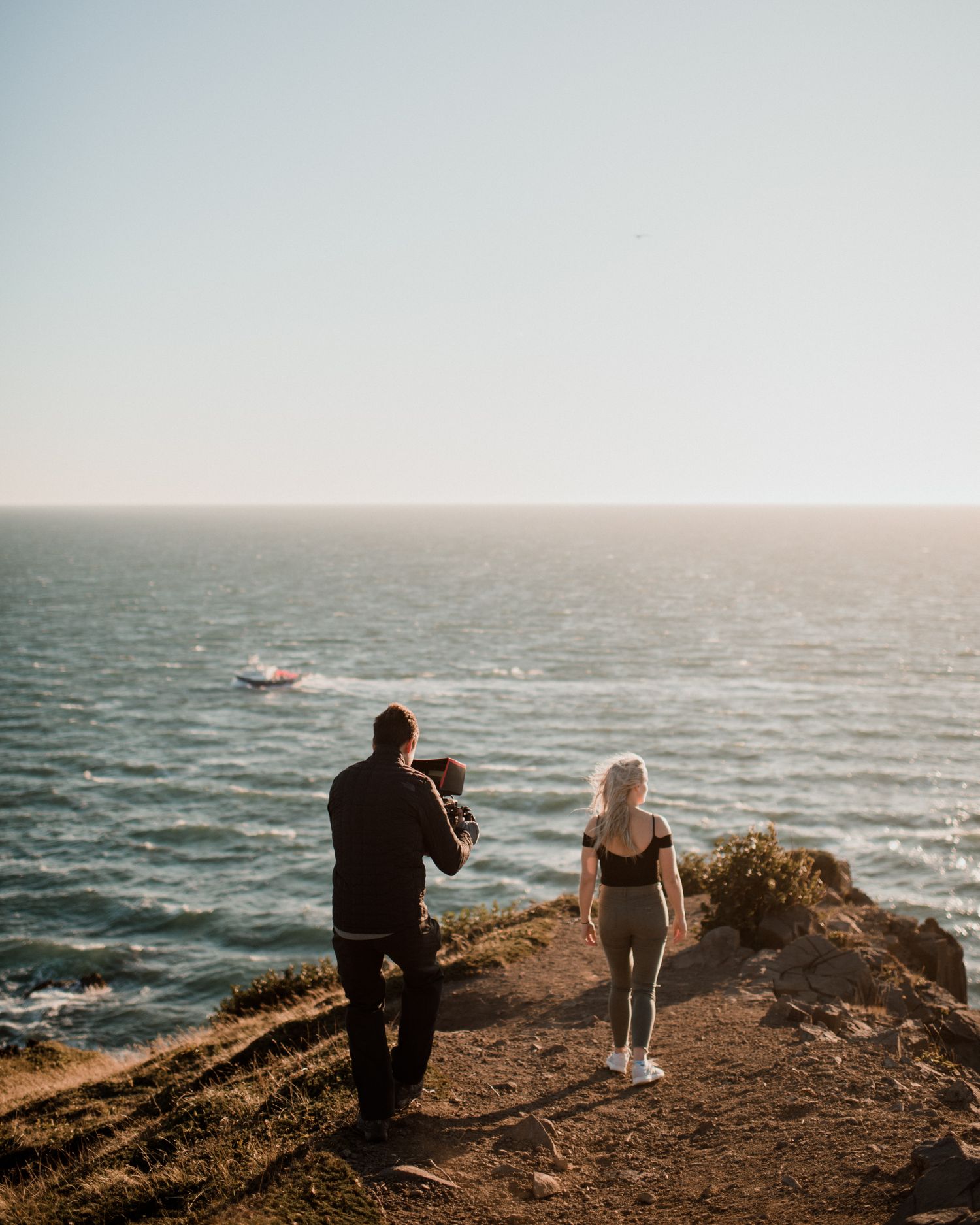 A couple with a baby stands on a coastal cliff overlooking the ocean with a boat visible in the distance.