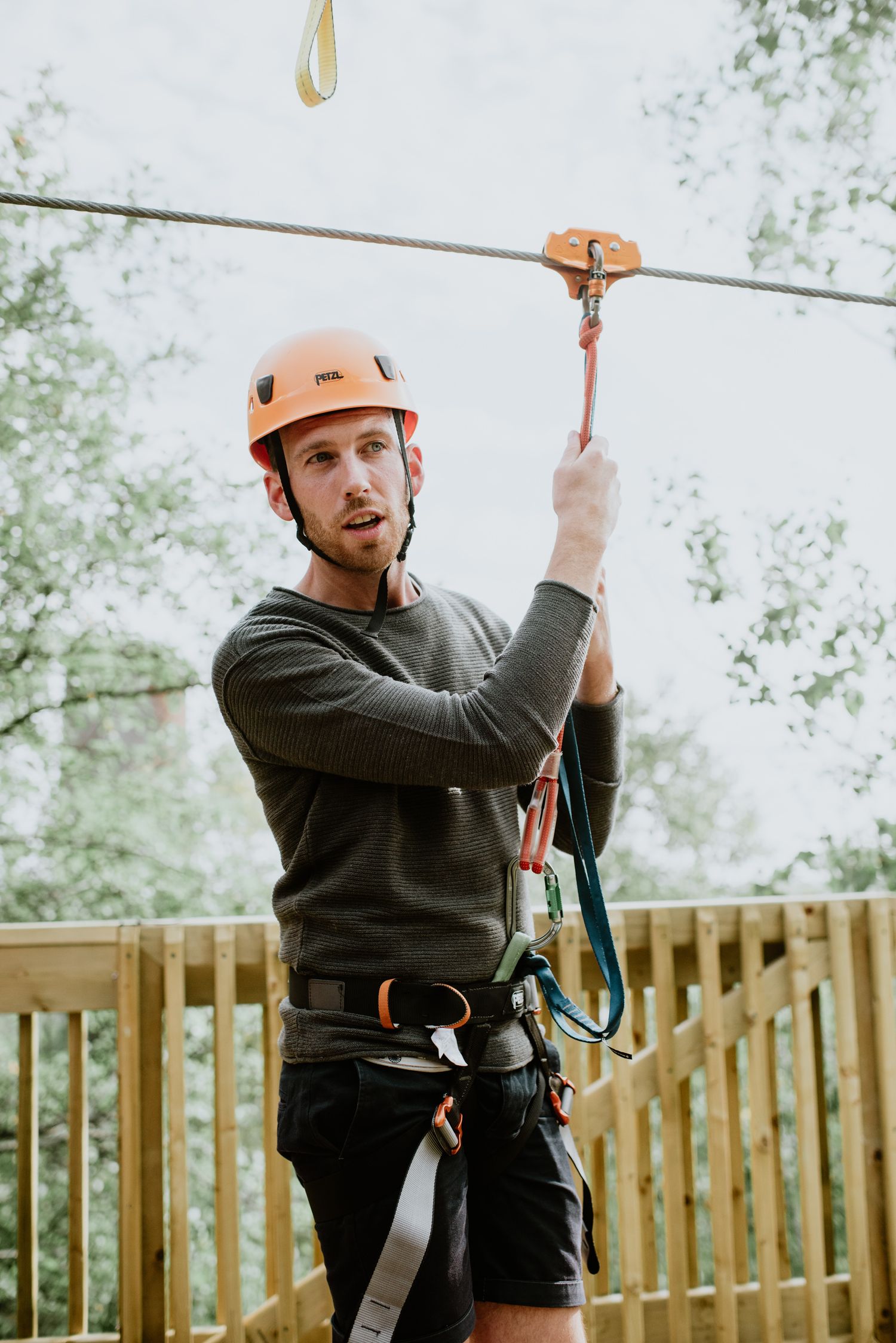 Man wearing orange helmet and harness on a zip line adventure course surrounded by trees.