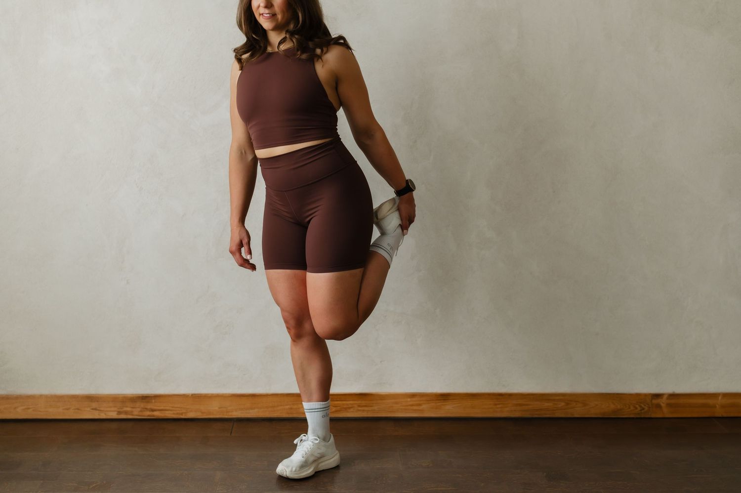 Athletic woman in brown workout set performs stretching exercises against neutral wall background.