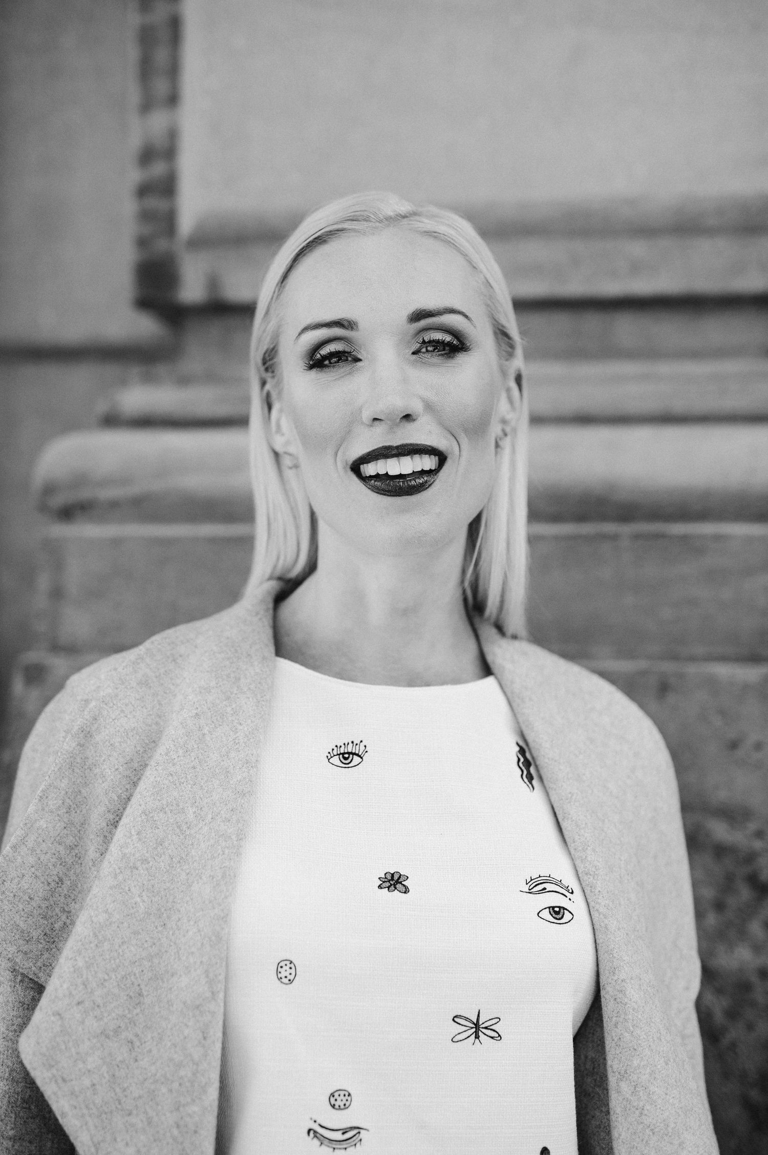 Smiling blonde woman in a light coat and printed dress posing outdoors in black and white.