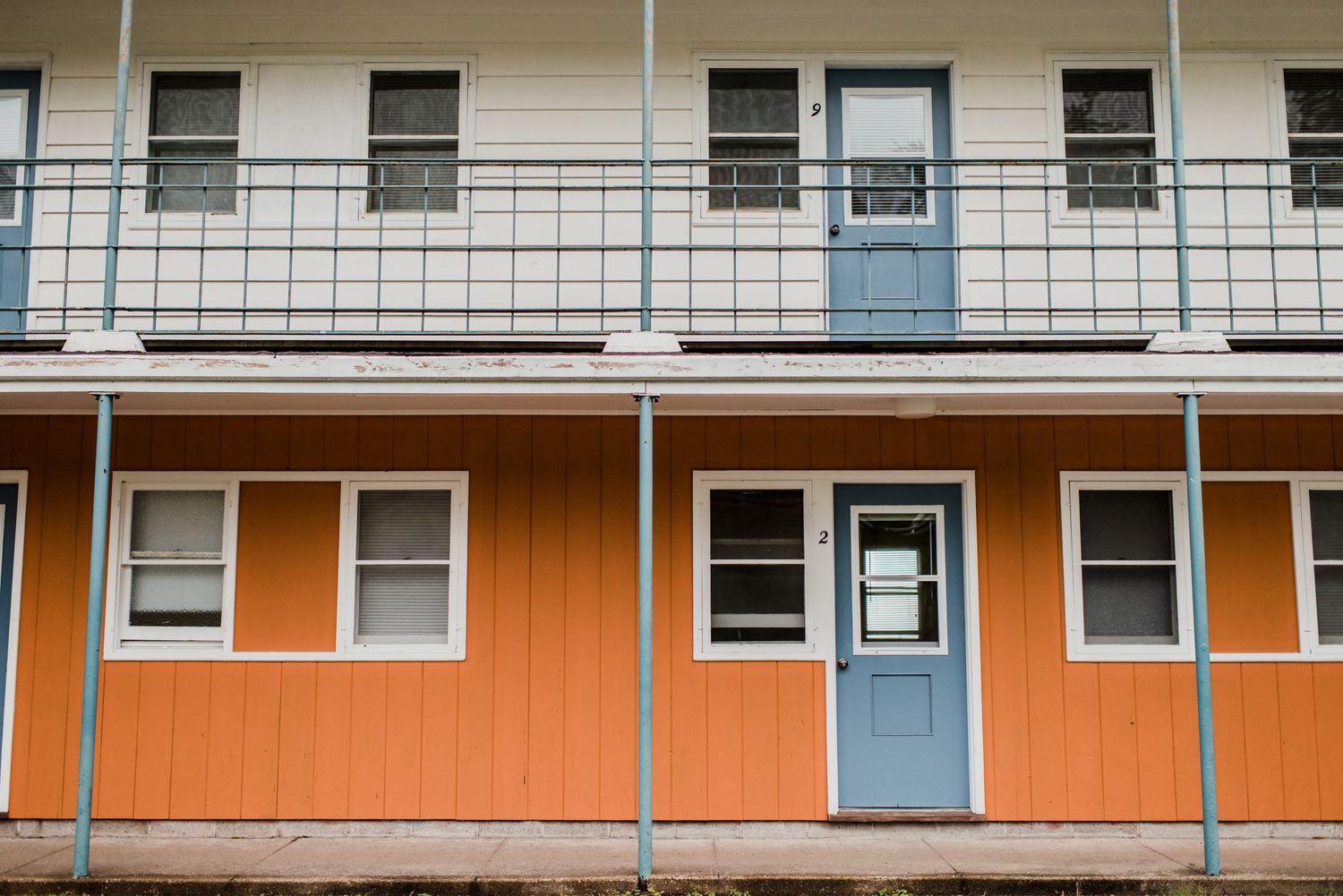 Two-story motel exterior with vibrant orange lower level, blue door, white windows, and white upper floor with balcony railing.