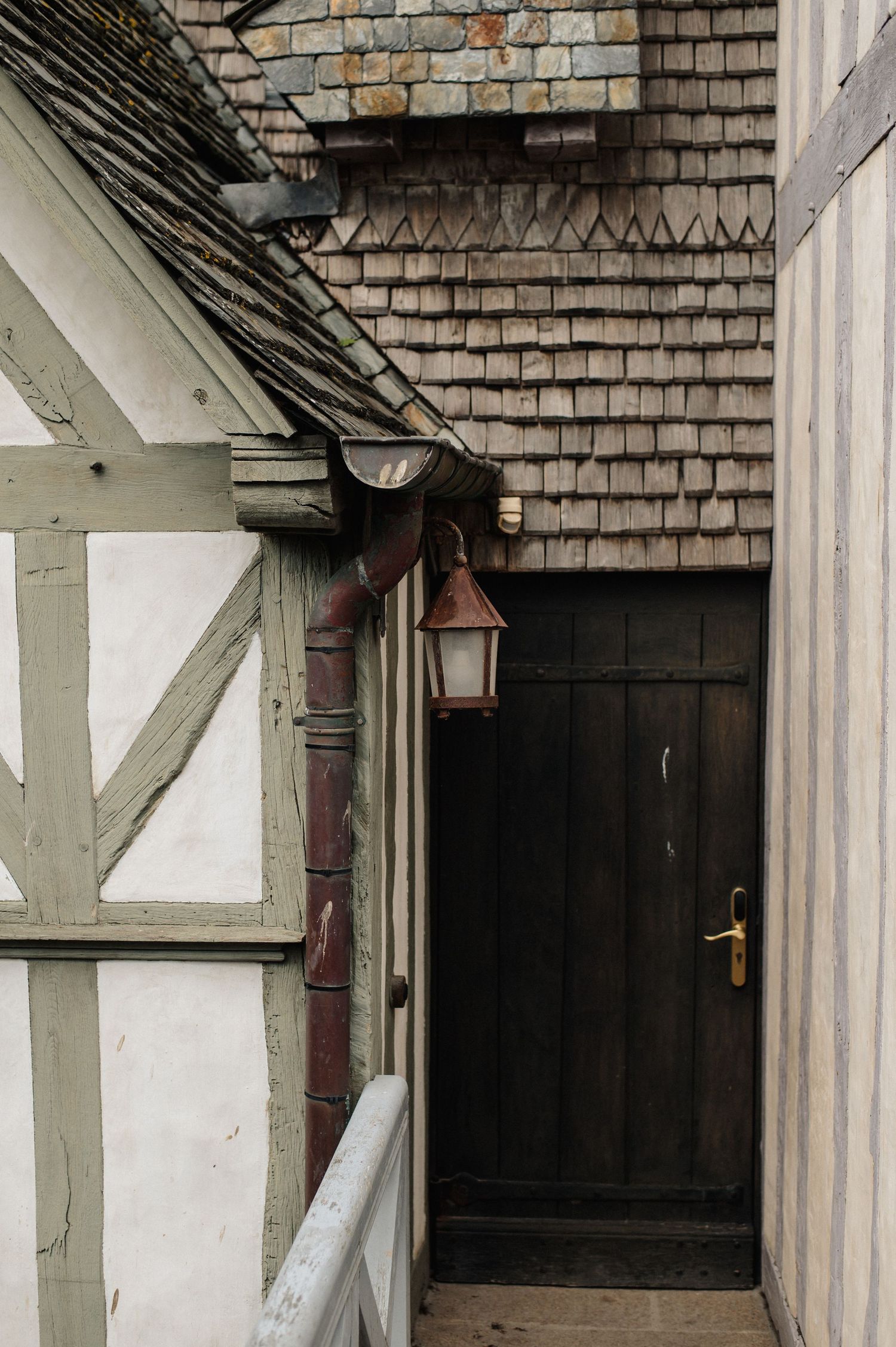 Medieval half-timbered building entrance with dark wooden door, copper drainpipe, and vintage lantern under wooden shingle roof.