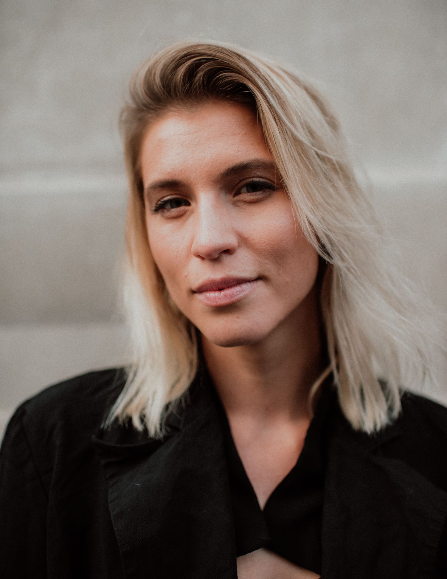 Young blonde woman with shoulder-length hair wearing a black jacket, posing outdoors against a stone wall.