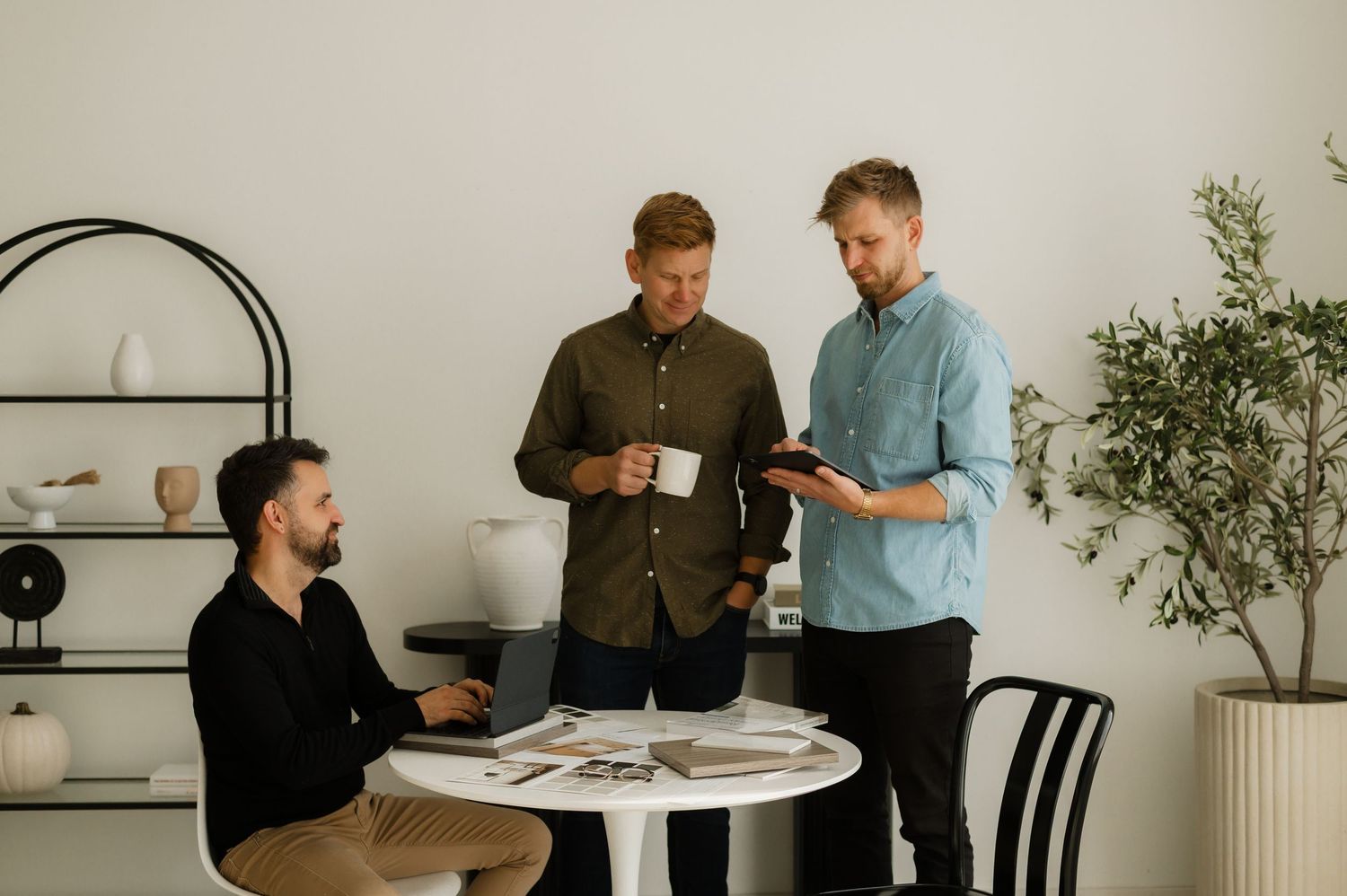 Three professional men collaborating in a modern office, reviewing documents and using a tablet at a white table.