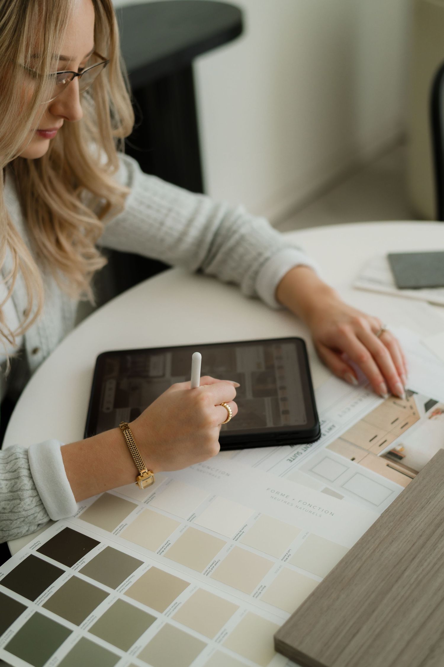 Blonde woman in glasses reviewing color swatches and floor plans on a tablet with stylus at a design desk.
