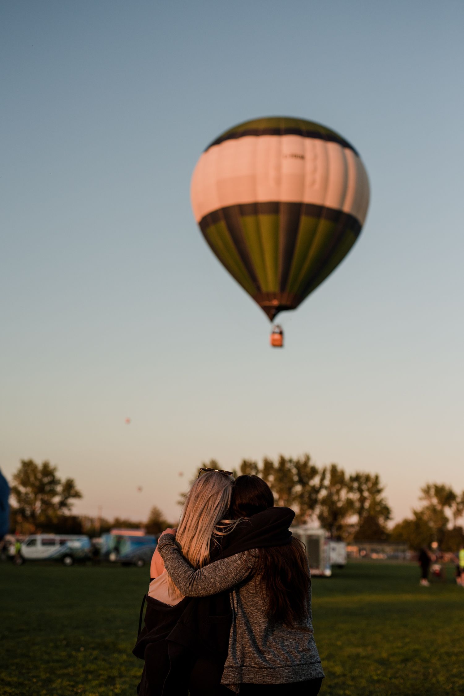Two people embracing while watching a green and white hot air balloon float in a clear evening sky over a grassy field.