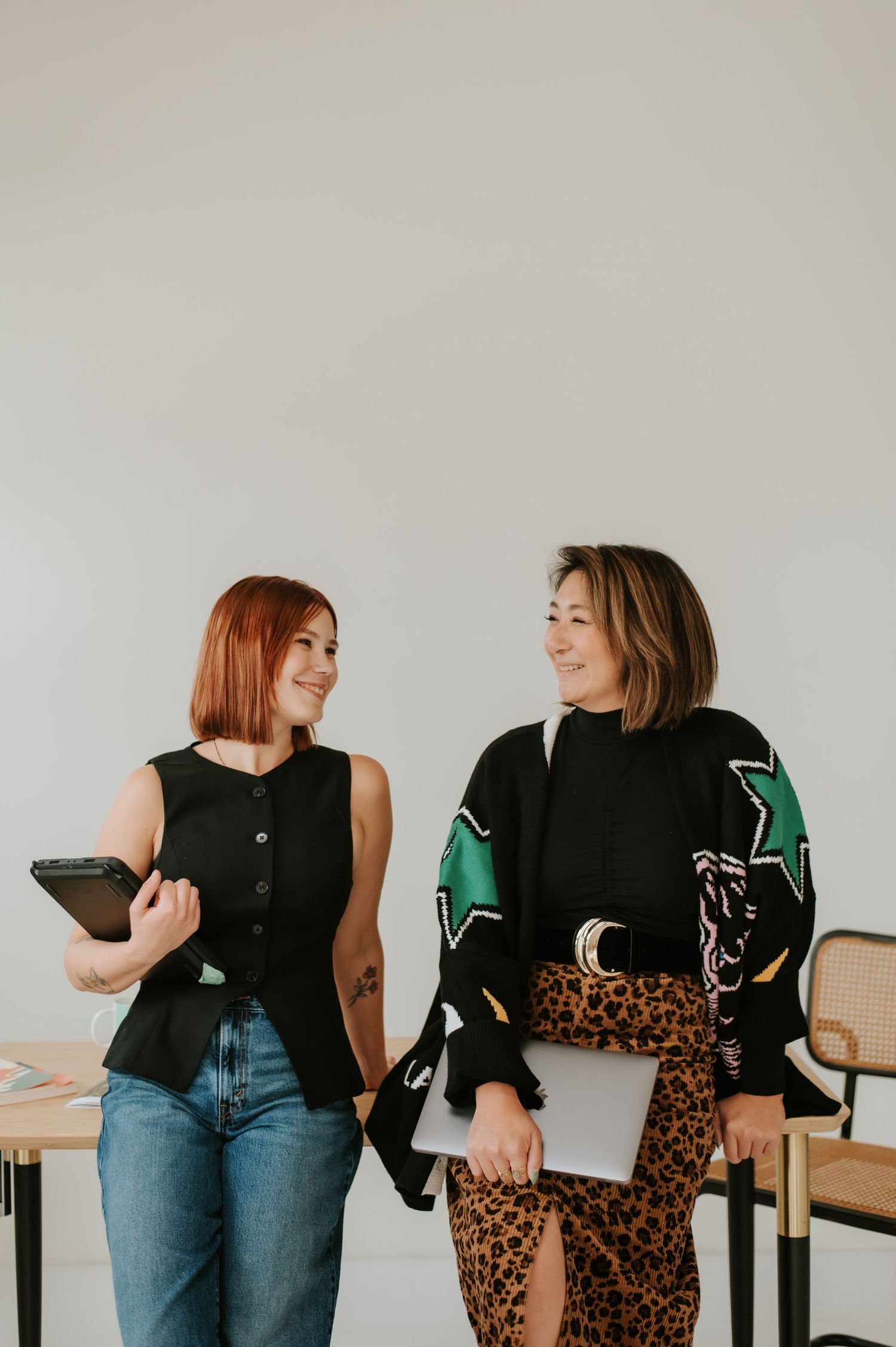 Two women in black tops and denim laugh and chat while sitting casually on chairs in a minimalist setting.
