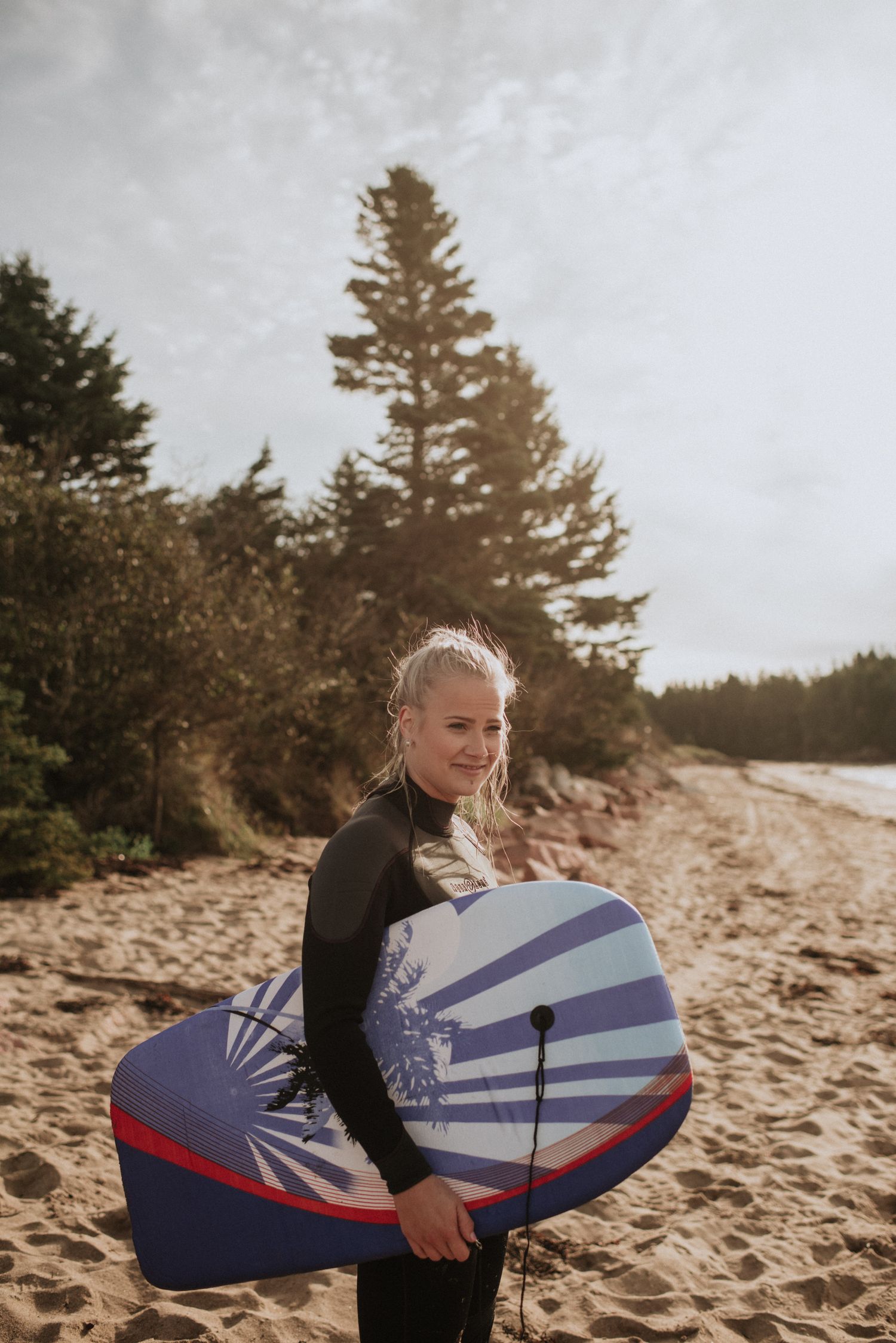 Young woman in wetsuit smiling on sandy beach, holding a blue and red bodyboard with trees in background.