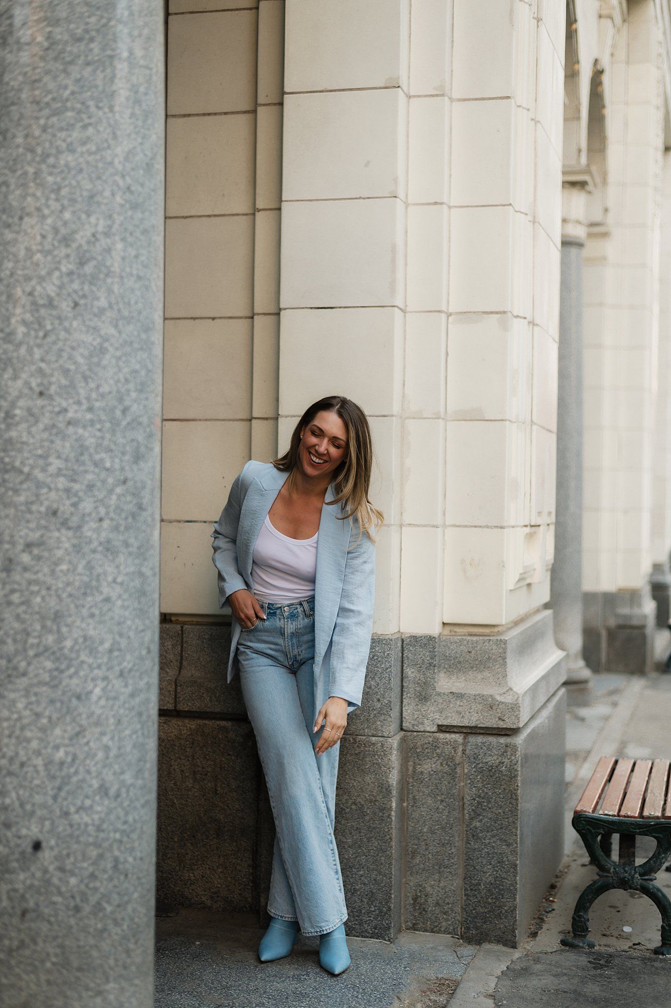 Woman in light blue blazer and jeans leaning against a stone building column, smiling outdoors.