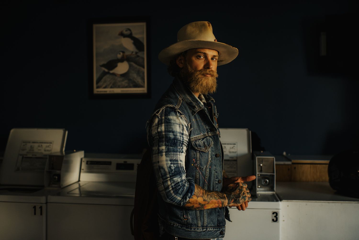 Bearded man in plaid shirt, denim vest, and cowboy hat stands in dimly lit room with vintage equipment.
