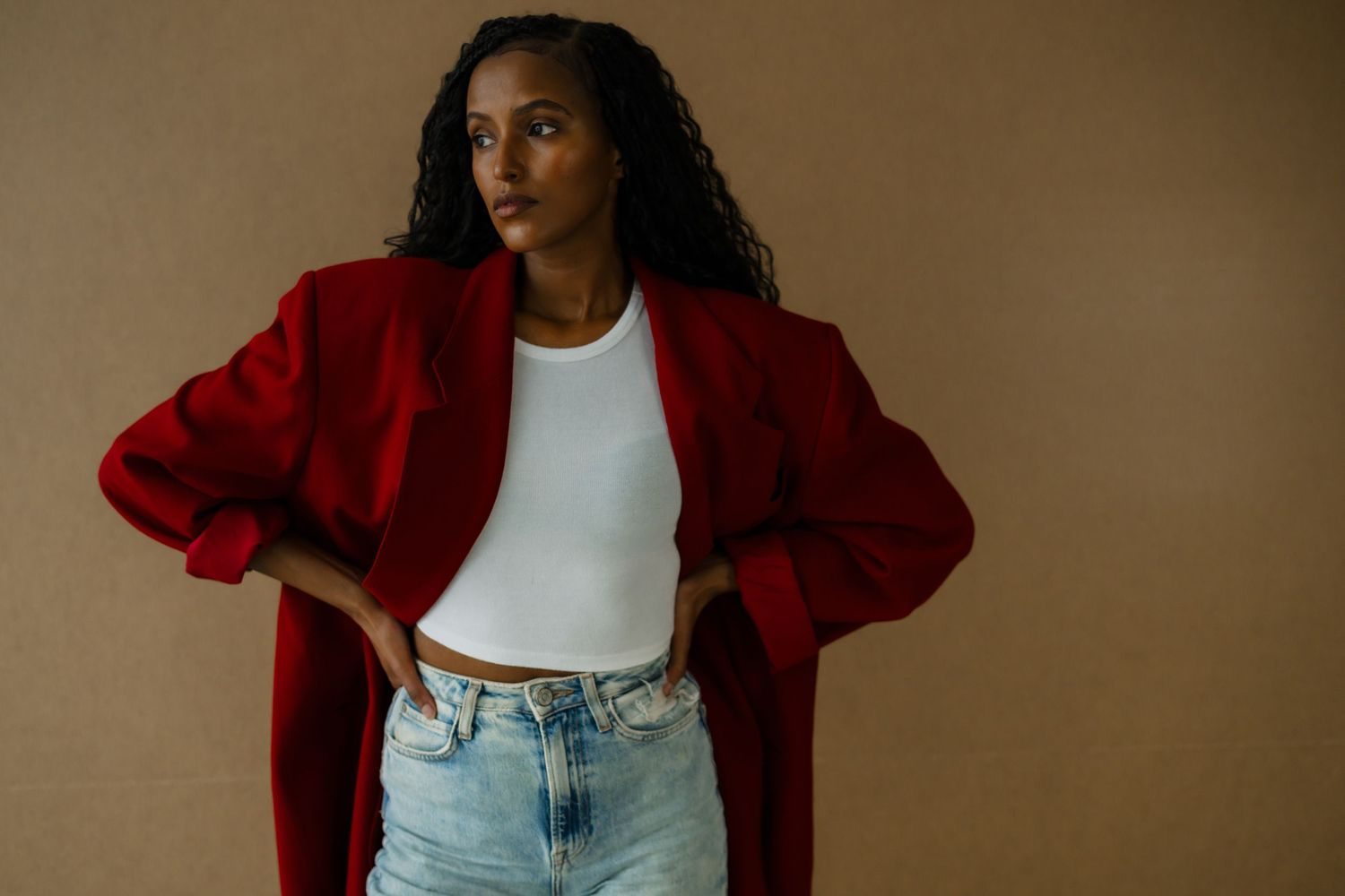 Woman in red blazer and white crop top with jeans posing confidently against a beige background.