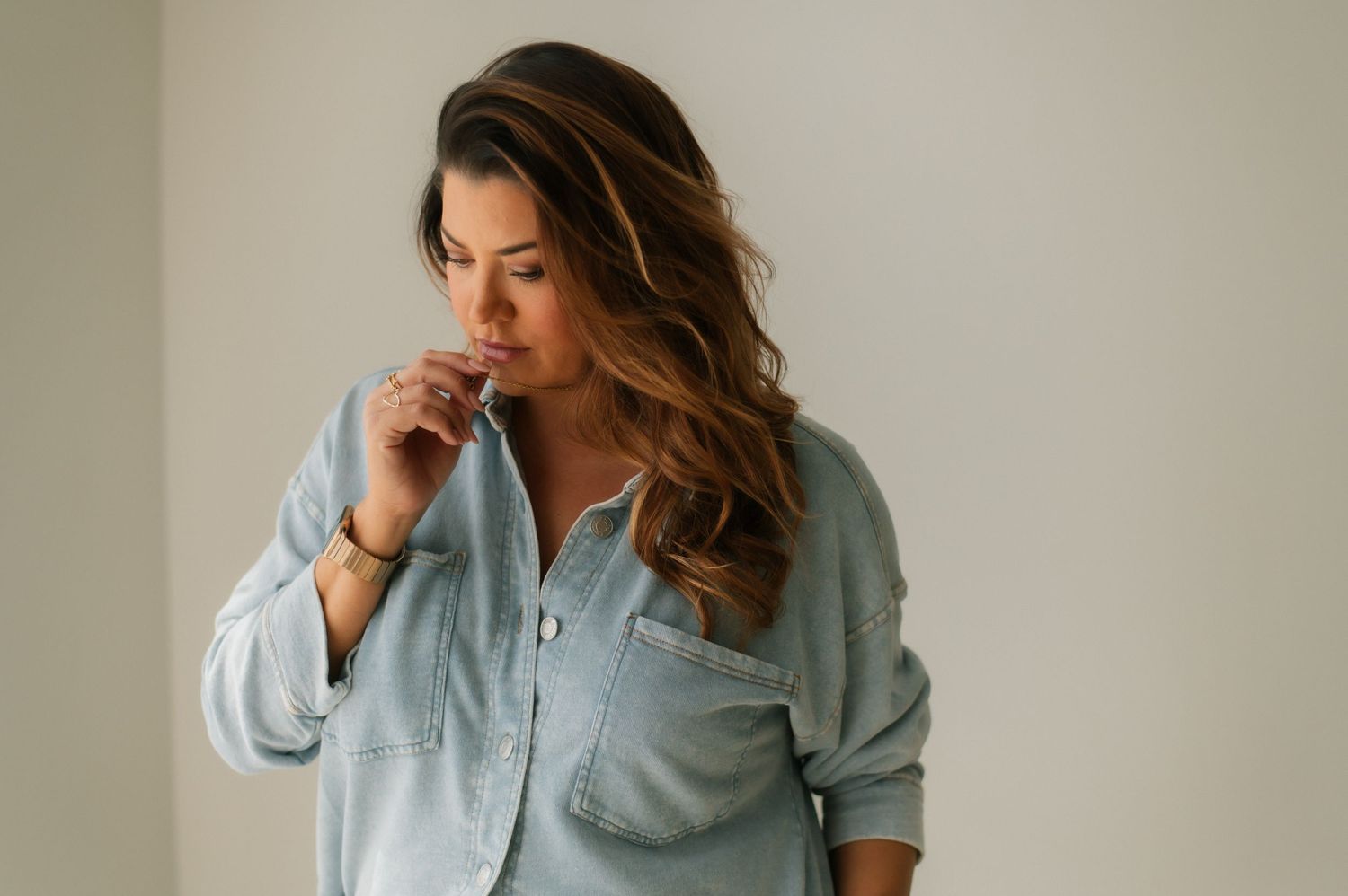 Person in casual light blue denim shirt poses thoughtfully against neutral wall with soft natural lighting.